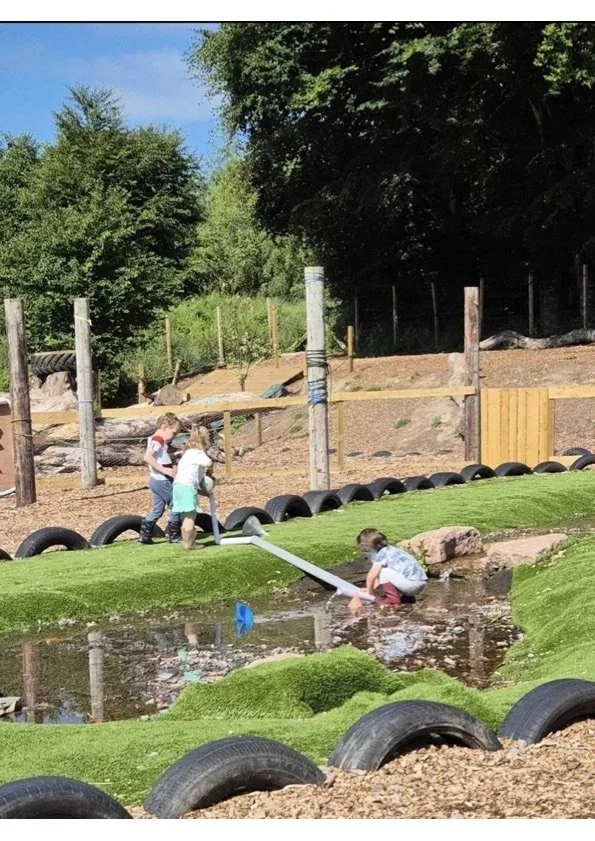 Three kids playing near a small artificial pond in a park, one of them using a small water pipe to fill a container, with a lush green landscape and wooden fencing in the background.
