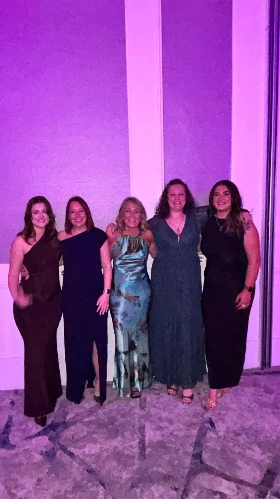 Five women in formal dresses posing together against a purple-lit wall.