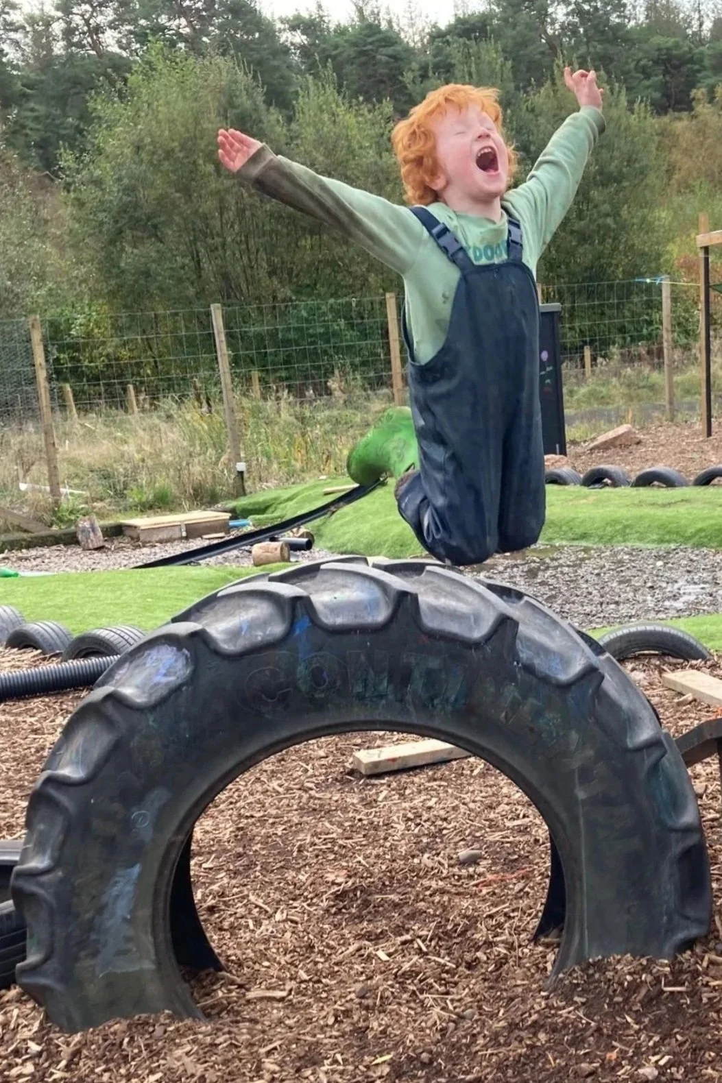 A young boy with red hair and green shirt jumping into the air with arms outstretched in a playground area, with large tires and wood chips on the ground, and trees and a fence in the background.