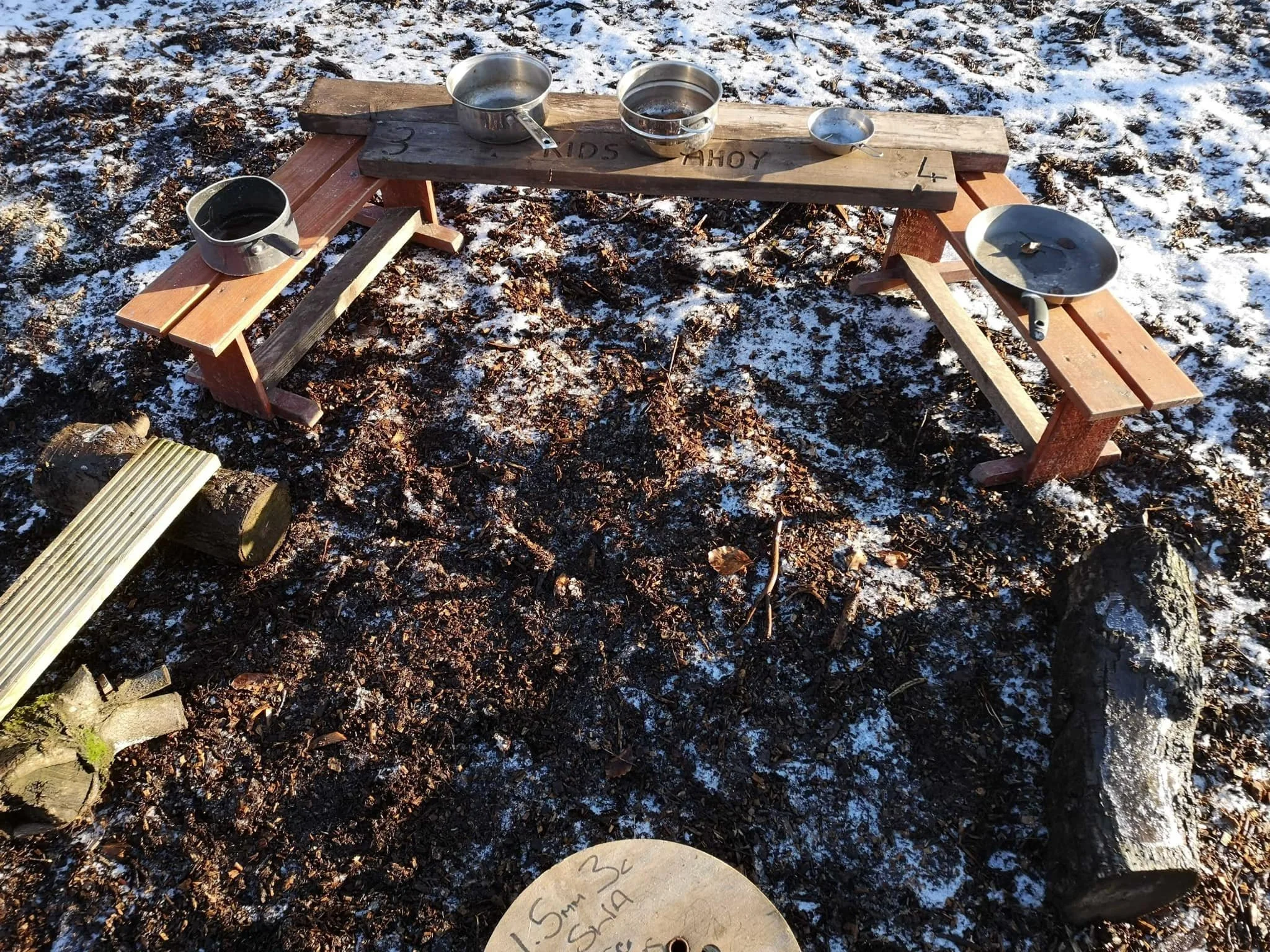 A small outdoor table with a wooden top and two attached benches, set on leaf-covered ground with snow. Metallic bowls and a cup are placed on the table, some with water inside.