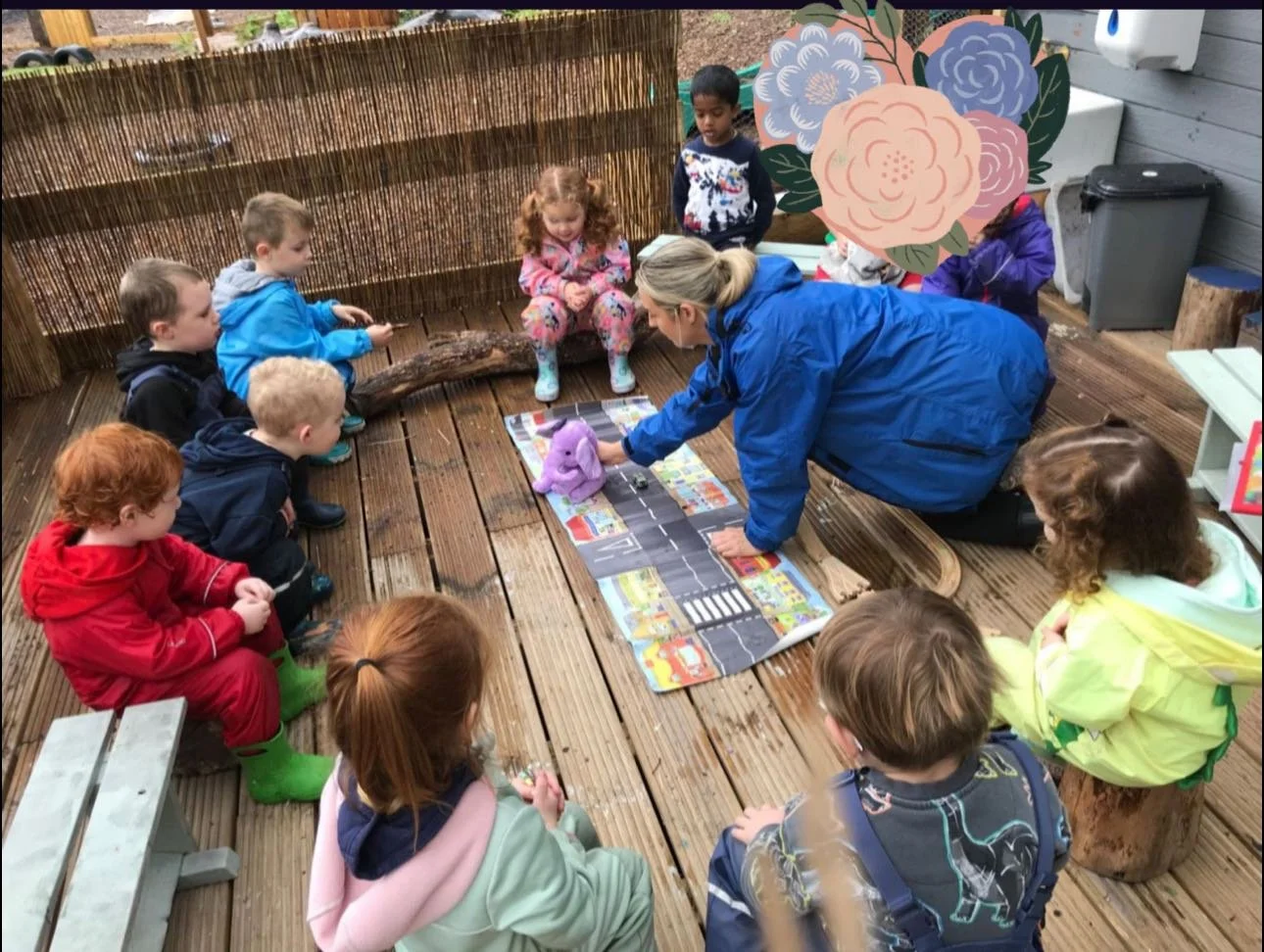 Group of young children sitting outdoors on a wooden deck around a teacher who is engaging them with a puppet and educational materials.
