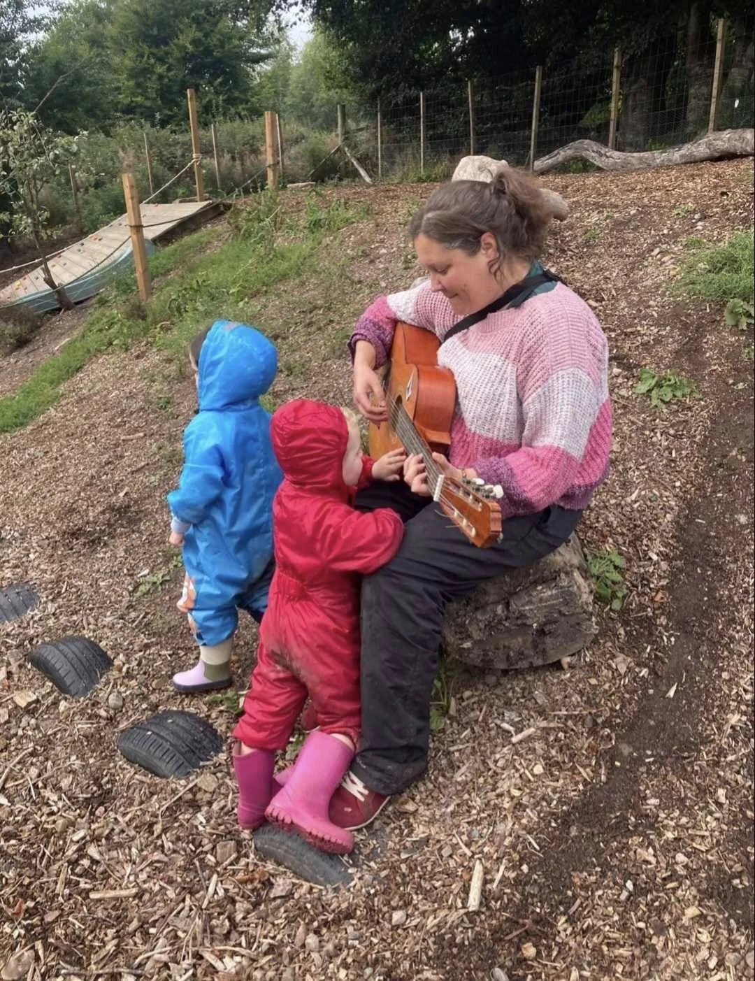 A woman is sitting on a tree stump playing a guitar outdoors, while two children in colorful raincoats stand nearby, listening. The setting is a natural, wooded area with a sloped ground, fencing, and plants.