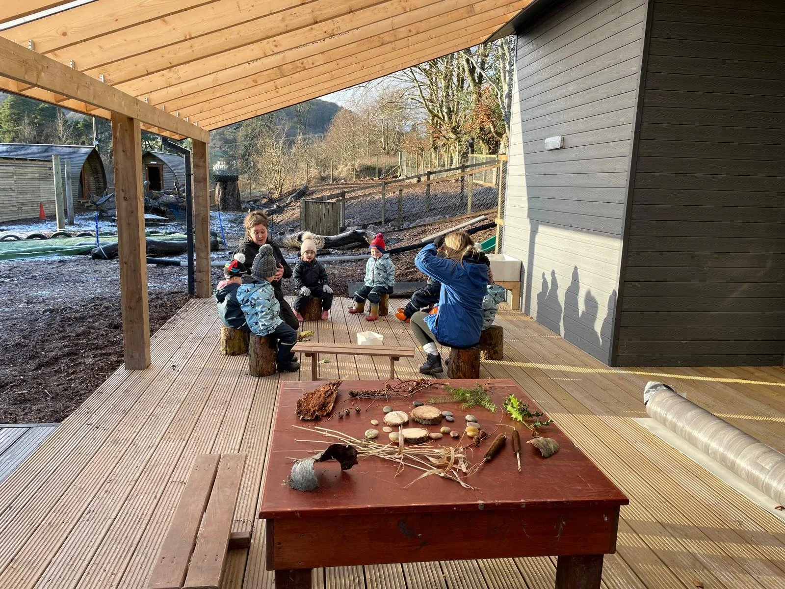 Children and an adult sitting on wooden stumps under a wooden roof outdoors, with natural scenery in the background. A table in the foreground displays natural objects like rocks, sticks, and pinecones.