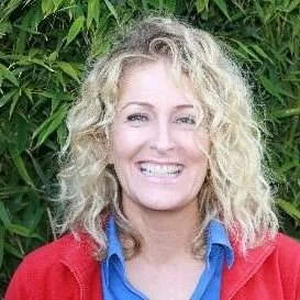 A woman with curly blonde hair smiling outdoors against green foliage.