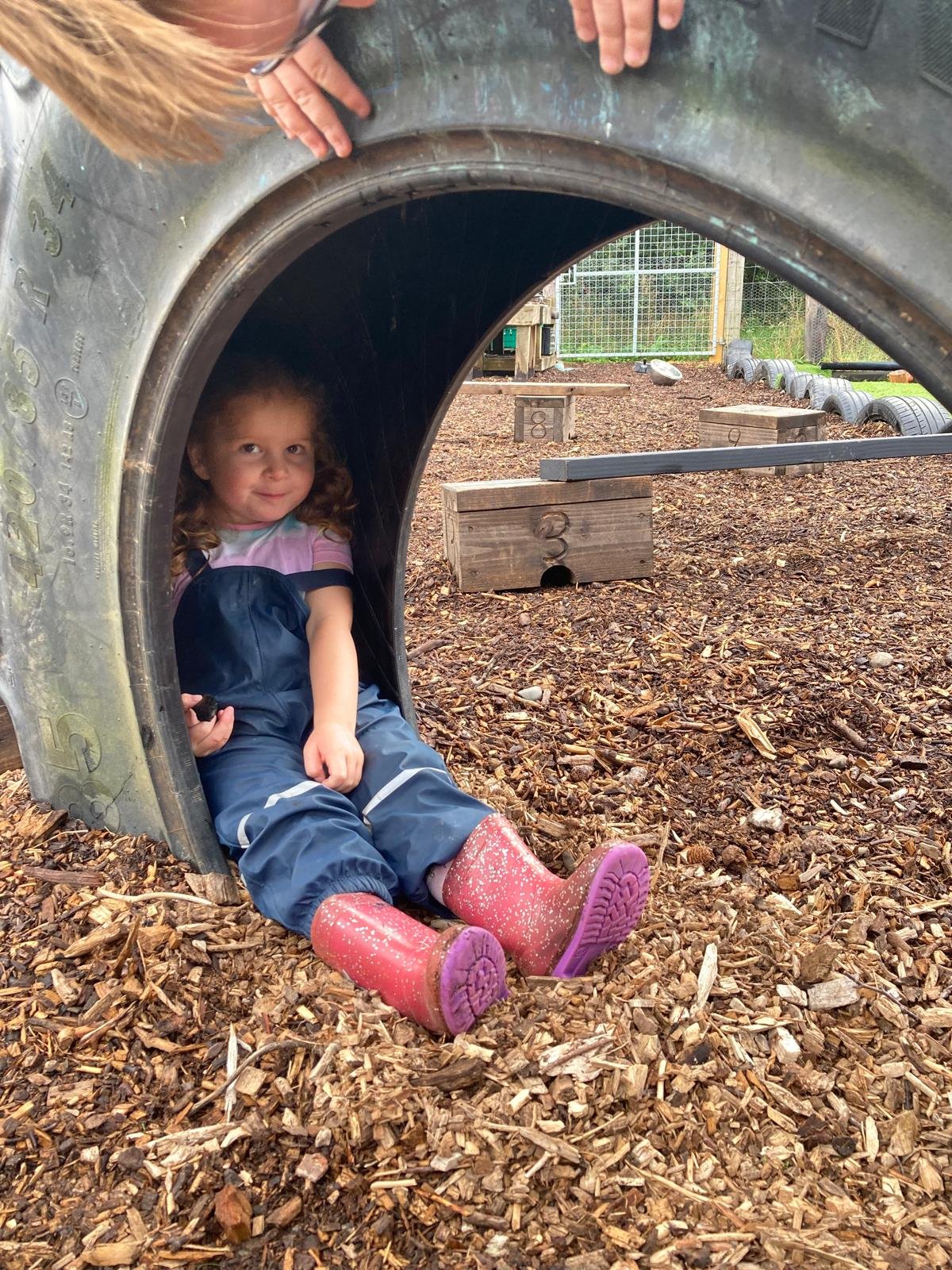 Child sitting inside a large, metallic, tunnel-shaped play structure at an outdoor playground, with wood chip ground covering, wooden obstacle blocks marked with numbers, and a chain-link fence in the background.