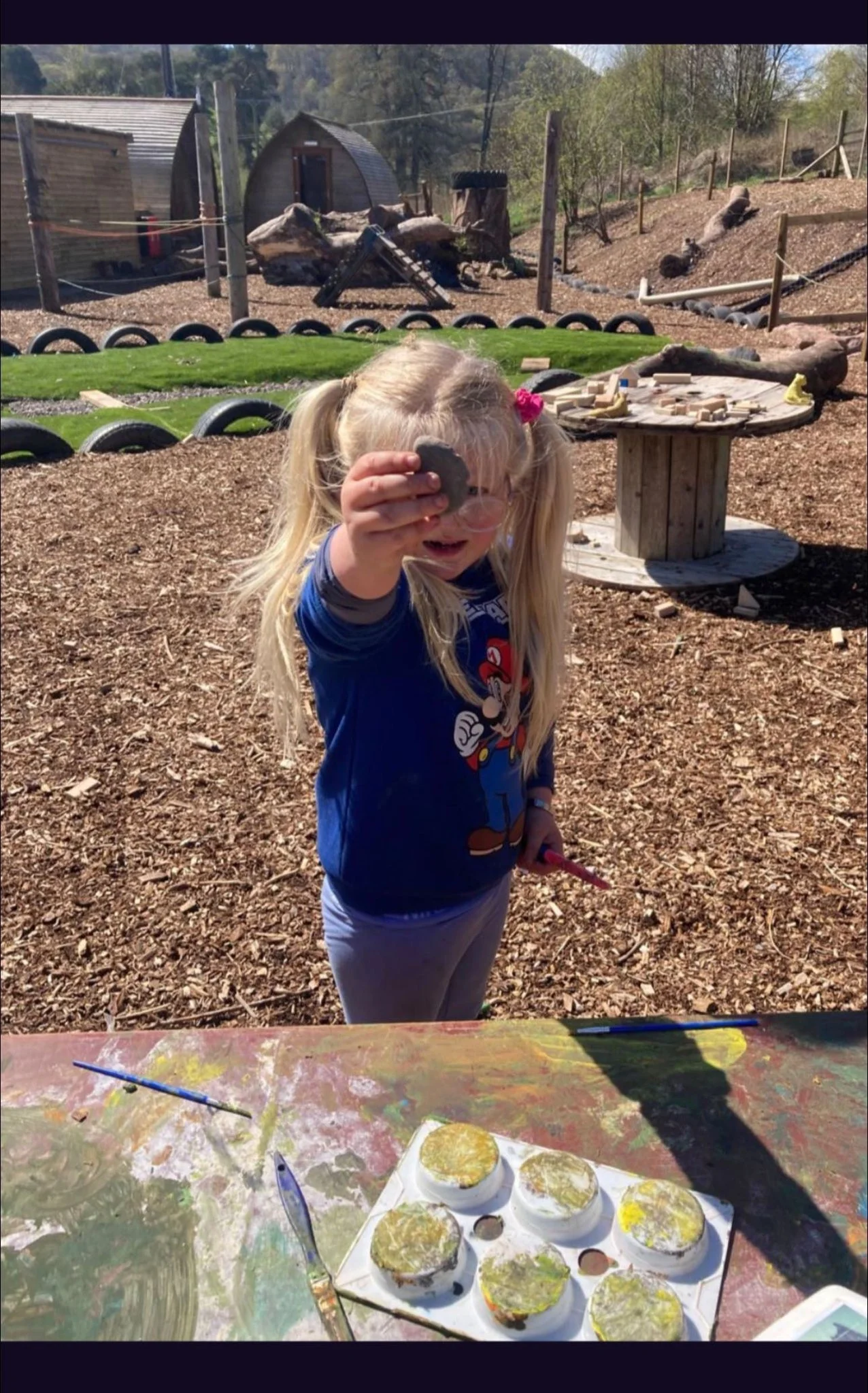 A young girl with blonde hair in pigtails holding a painted rock in an outdoor art studio with a colorful table, paints, and brushes, surrounded by a natural landscape with trees, fencing, and a wooden shed.