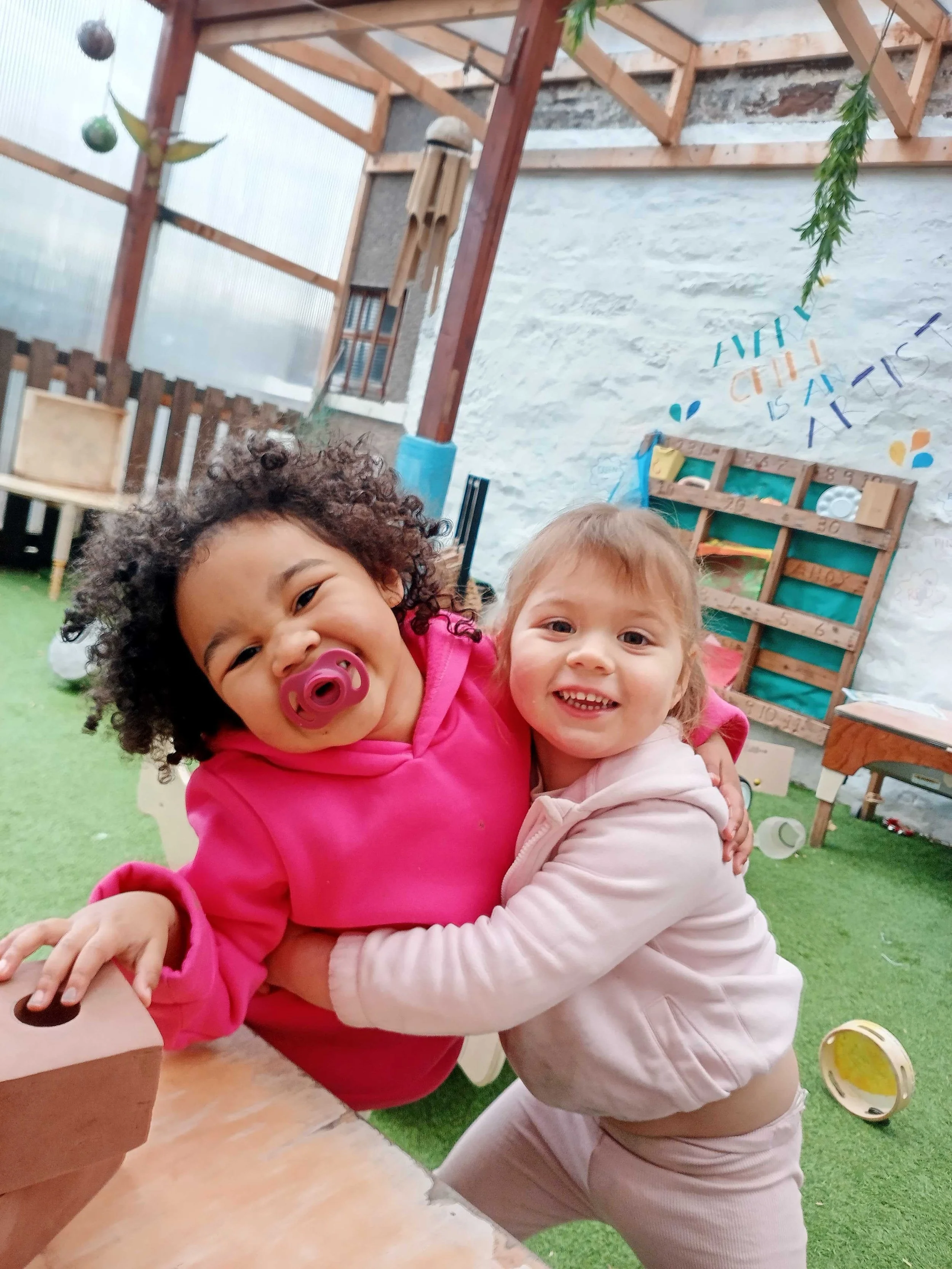 Two young children hugging and smiling in a play area with a green carpet, wooden furniture, and a decorated wall in the background.