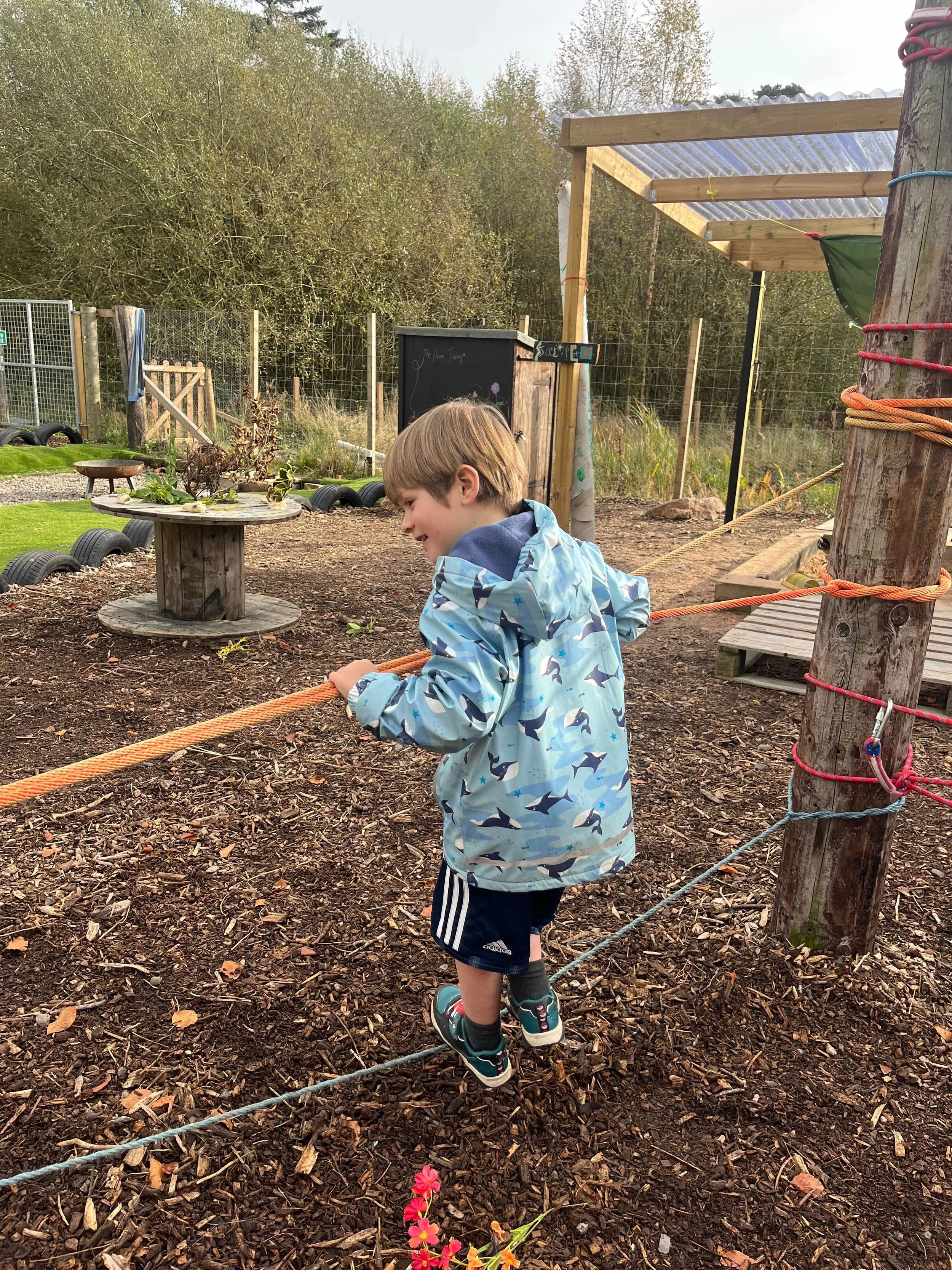 A young boy in a blue jacket with shark print walking on a rope in an outdoor playground area surrounded by mulch, with a large wooden pole and a rustic fence in the background.