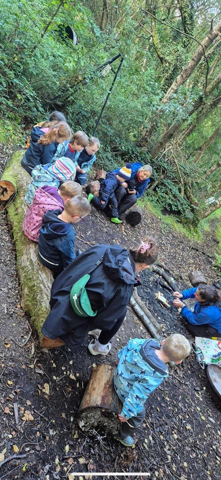 Adults and children sitting and standing around a small outdoor fire pit in a wooded area, engaging in a nature educational activity.