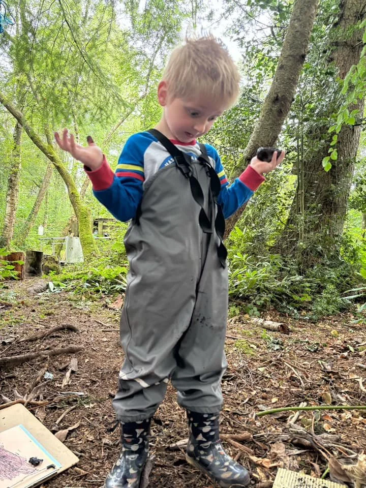 A young boy with a confused or surprised expression, wearing gray waterproof overalls, patterned rain boots, and a colorful jacket, standing in a wooded outdoor area with trees and greenery.