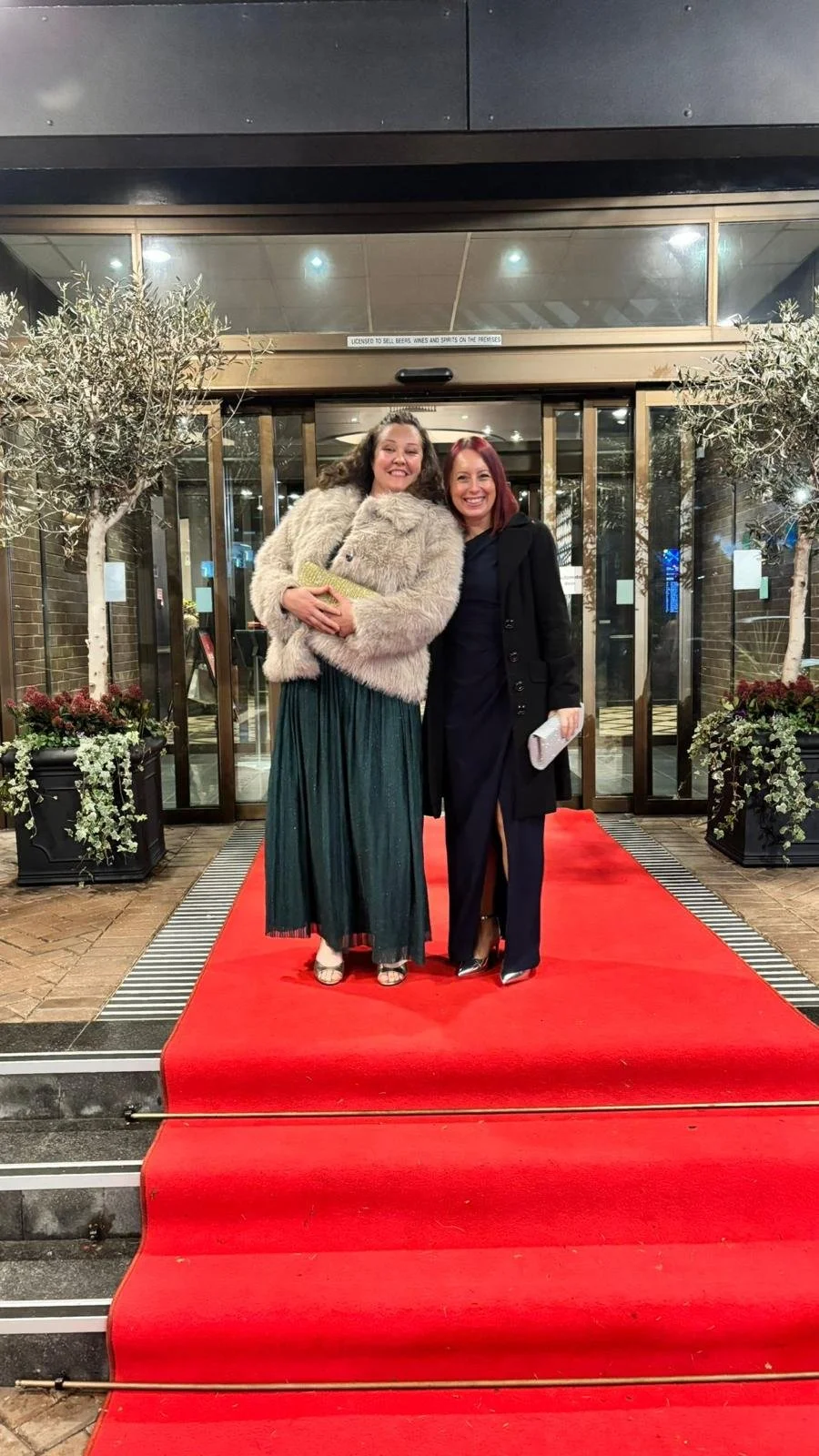 Two women standing on a red carpet outside a building, smiling and posing for the photo.