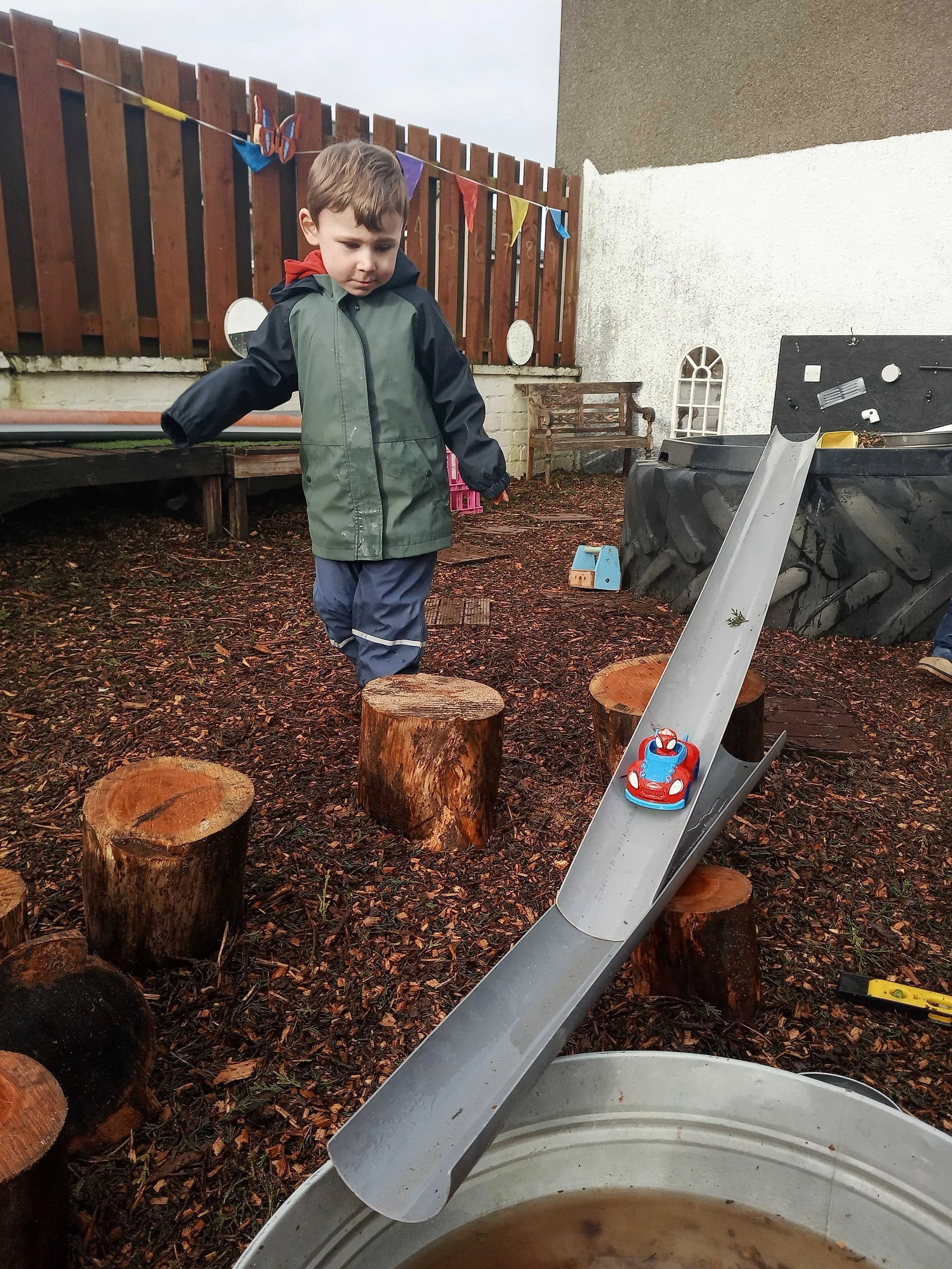 A young boy in a green jacket and gray pants playing outside near a slide and logs with a wooden fence and colorful flags in the background.