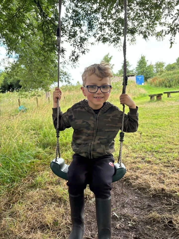 A young boy with glasses, wearing a camouflage jacket and black pants, is sitting on a swing in a grassy outdoor area under a tree, smiling at the camera.