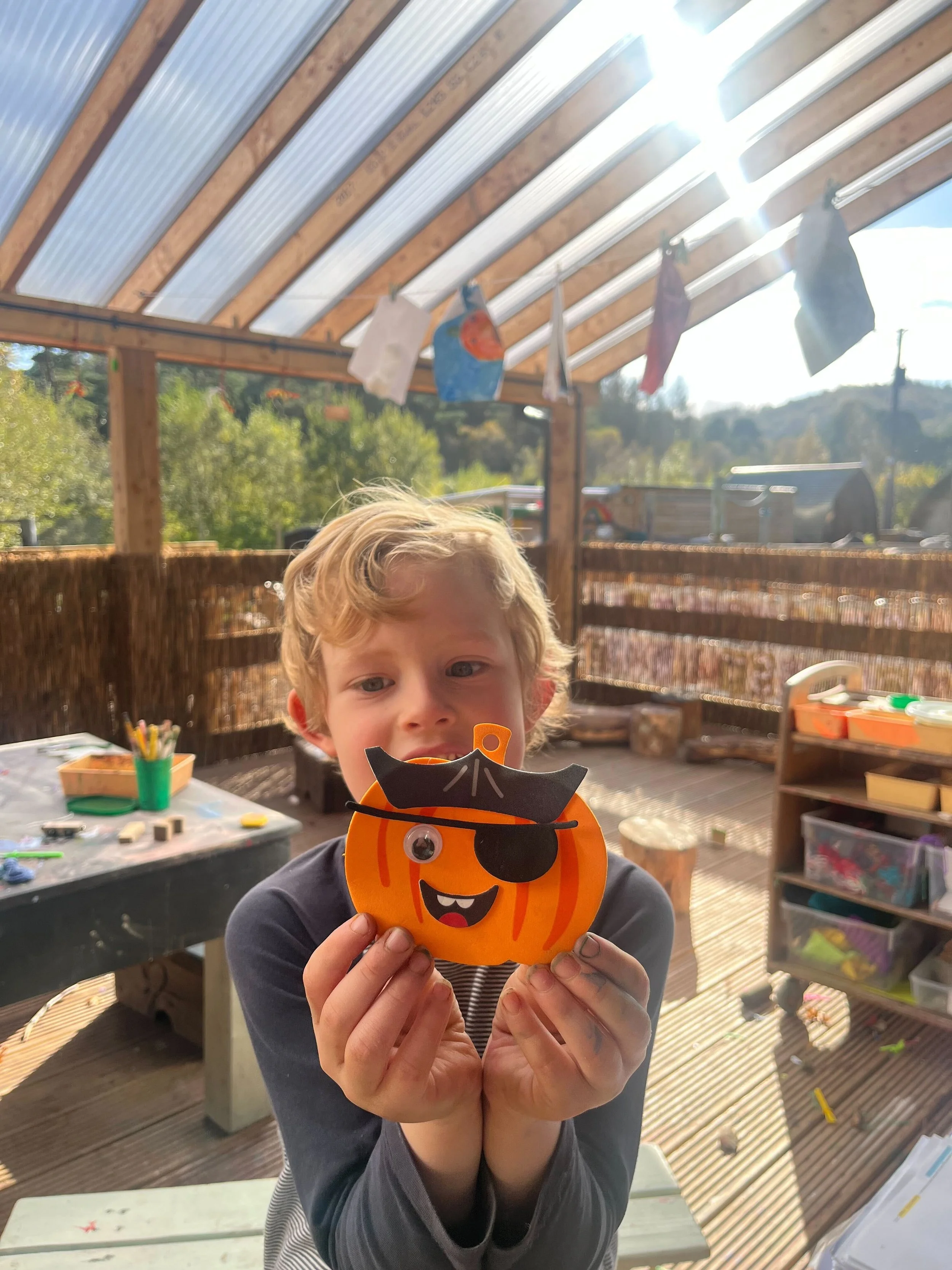 A young boy with blonde hair holds a paper pumpkin craft with a cartoonish face and an eyepatch, standing outdoors under a shed with solar panels and colorful laundry hanging. There are trees and a wooden fence in the background.