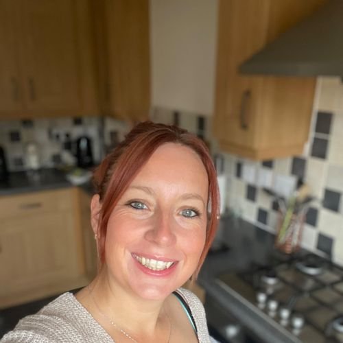 A woman with red hair smiling in a kitchen with wooden cabinets and a black and white tiled backsplash.