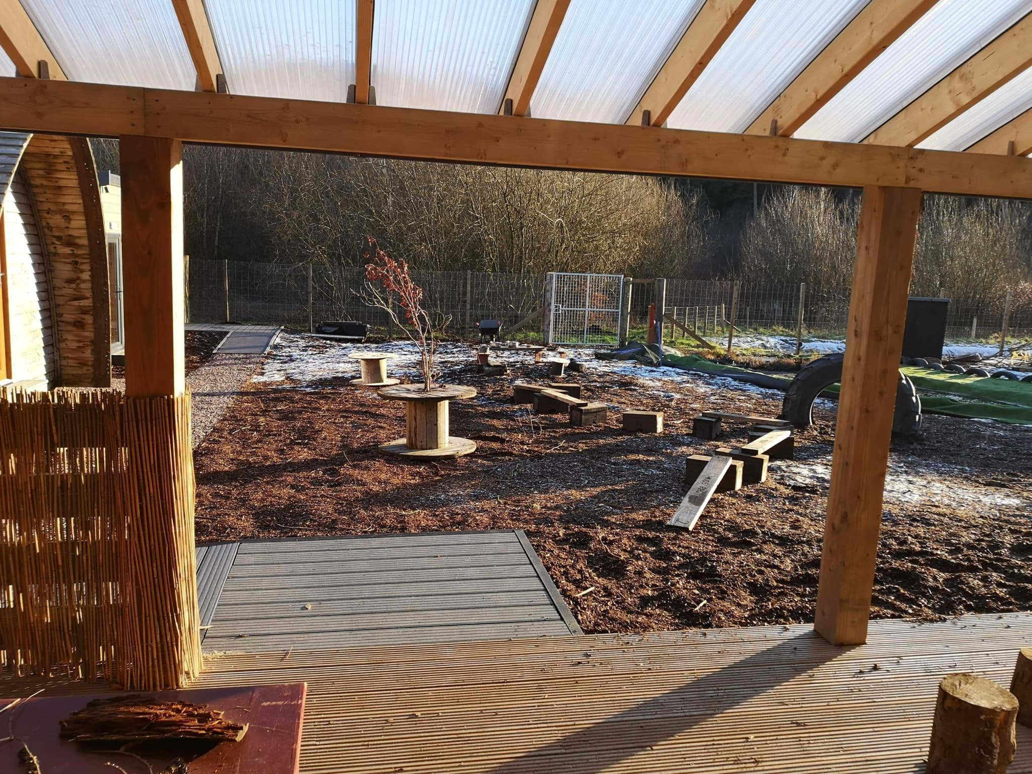 Construction site showing a wooden structure with beams and partially finished porch, outdoors with a fenced backyard and leafless trees.
