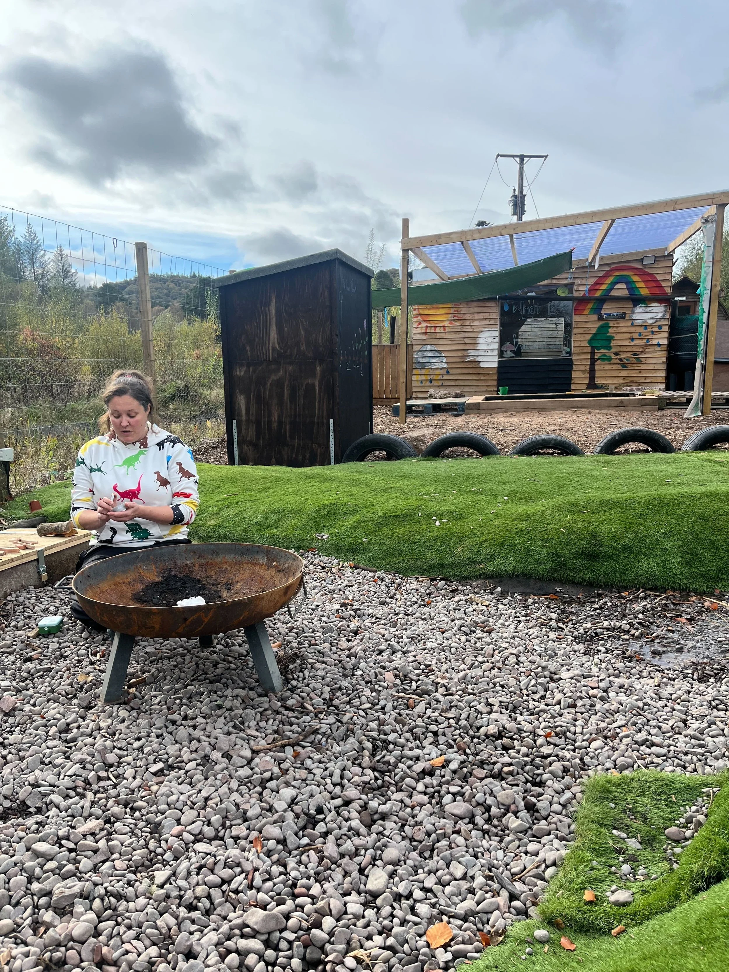A woman sitting outdoors on a rocky ground next to a large rusted fire pit, with a green artificial grass area, a black shed, and a colorful painted wooden playhouse with a rainbow, clouds, sun, trees, and rainbows painted on it. The sky is partly cloudy with dark clouds.