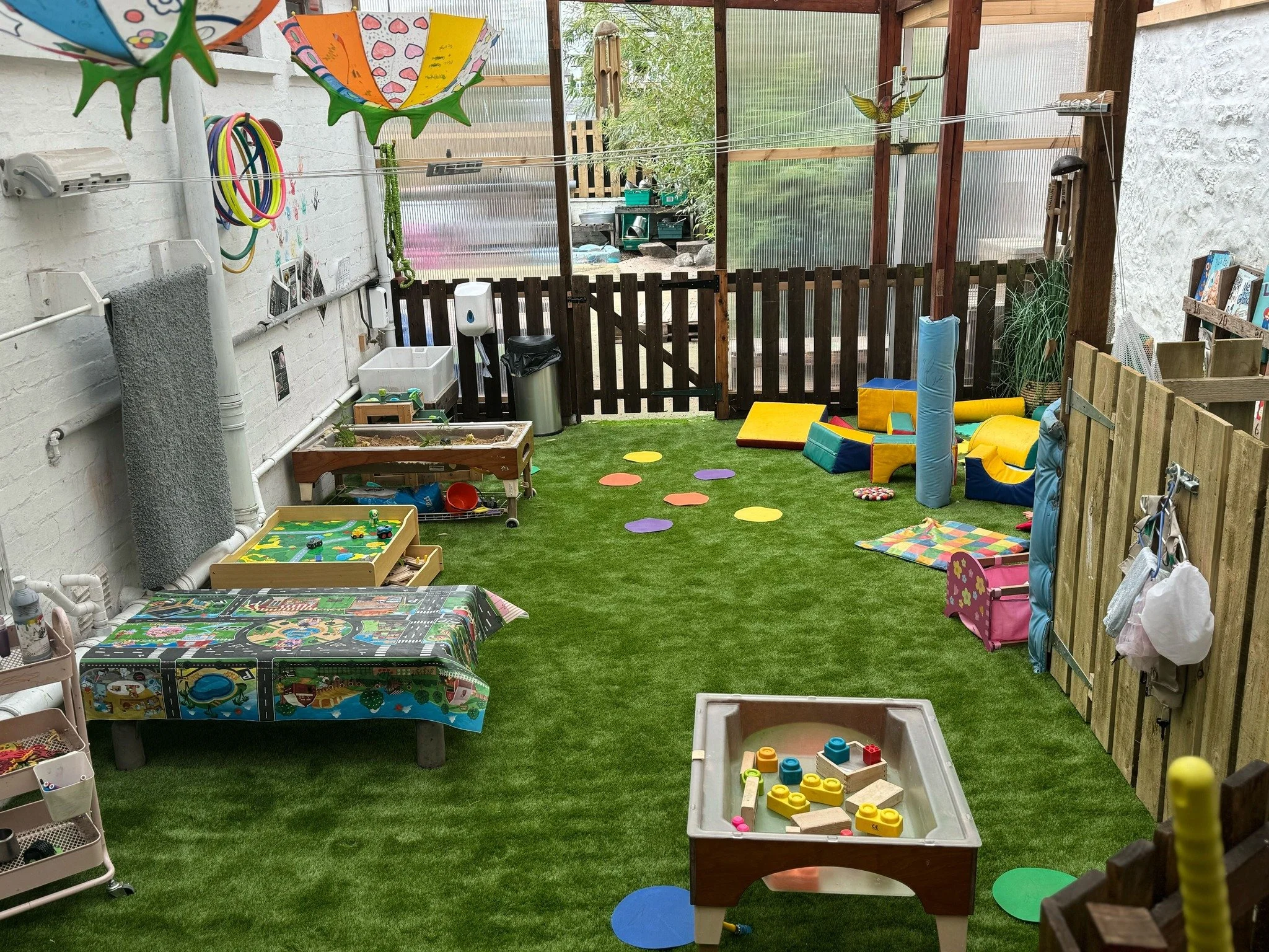 A colorful enclosed outdoor play area for children with various toys, play mats, and decorations on the walls and fence.