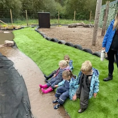 Four children sitting by a small pond, with one standing nearby, at an outdoor water play area with artificial grass and tire border.