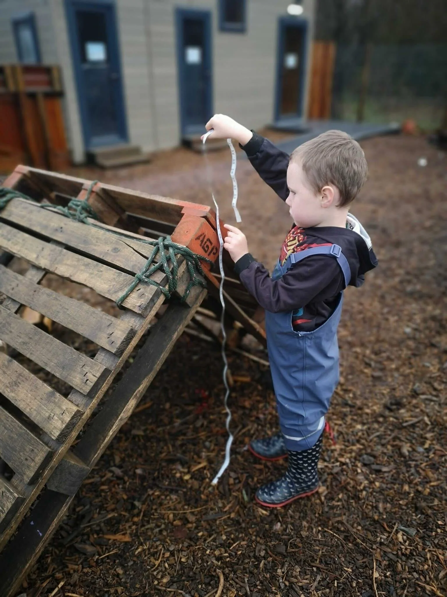 A young boy in a blue jacket and rain boots holds a piece of measuring tape, standing outdoors near a wooden pallet and a building with multiple small windows.
