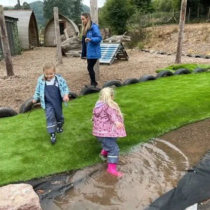 Two young children, a boy and a girl, walk on a small artificial grass hill near a muddy water area, with a woman in the background watching them, in an outdoor play area.
