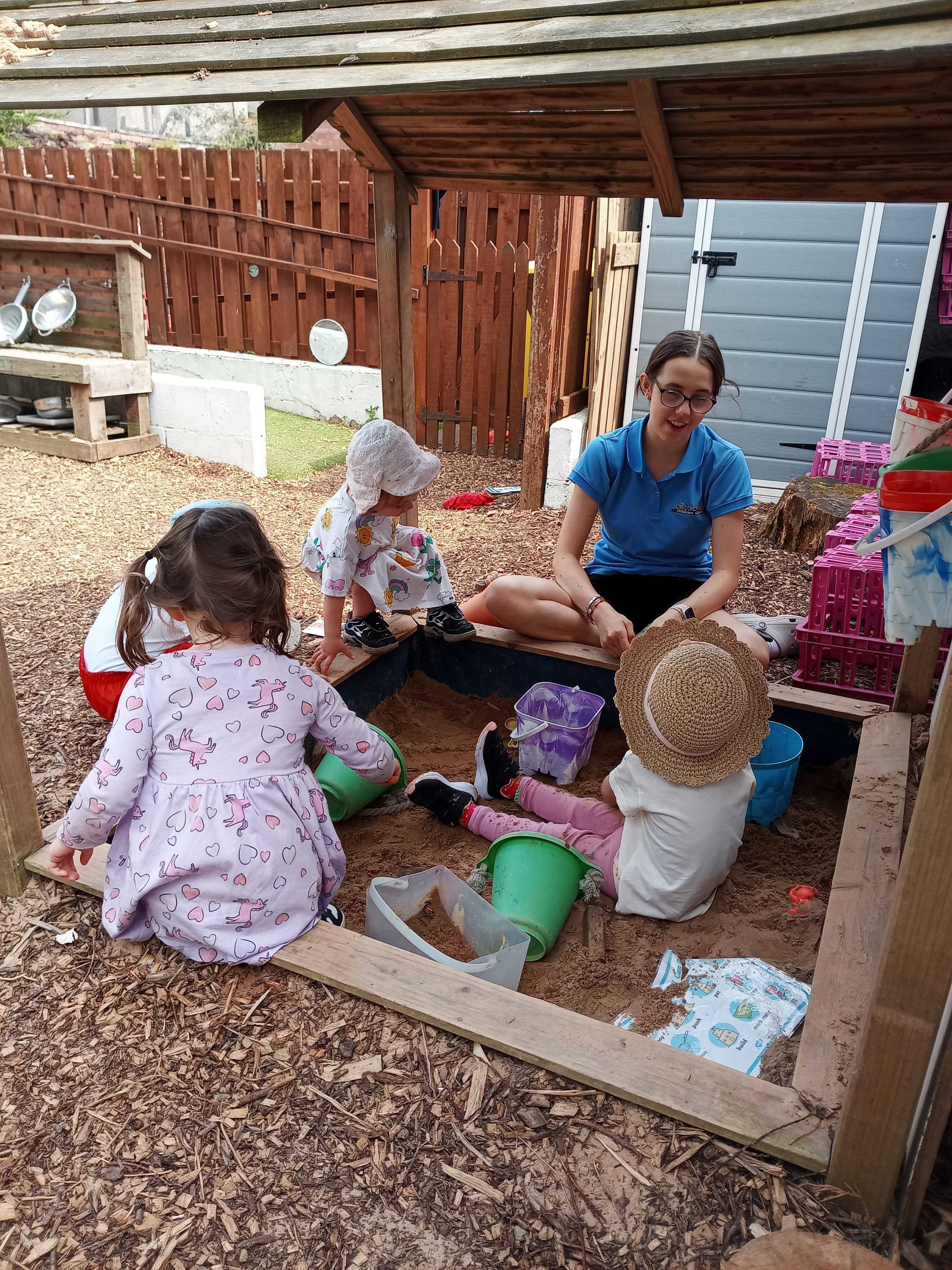 Children and an adult playing in a sandbox under a wooden shelter in a backyard, with a wooden fence, garden tools, and storage shed in the background.
