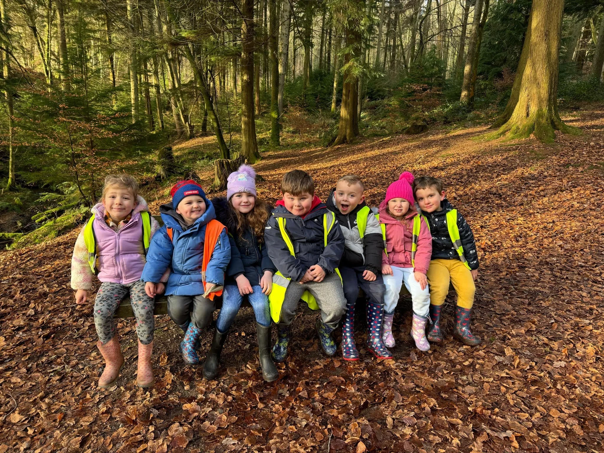 A group of eight young children standing in a line outdoors in a forest, dressed in colorful jackets and rain boots, with trees and fallen leaves in the background.
