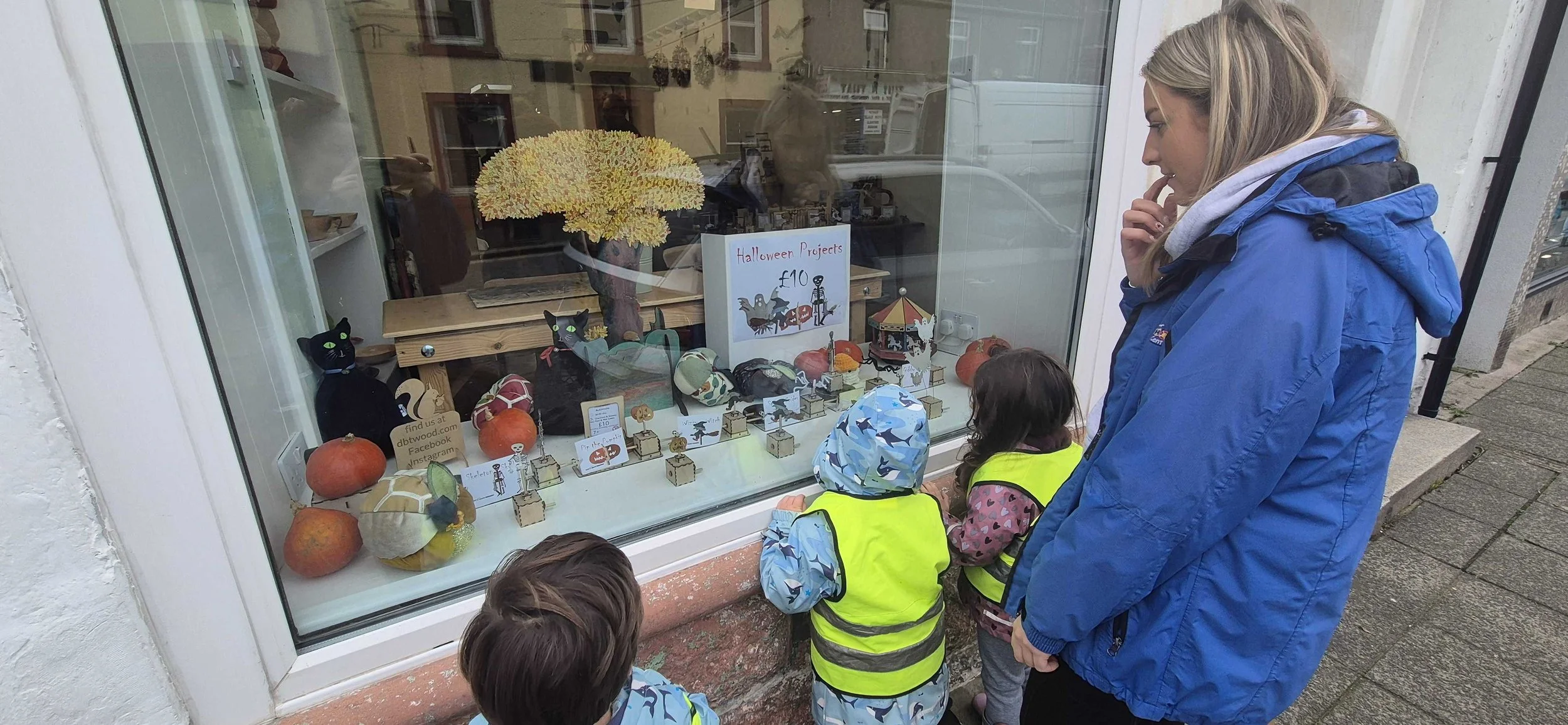 People looking at Halloween-themed crafts and decorations in a shop window display, including painted pumpkins, cat figures, and paper cutouts, with a sign indicating these are Halloween projects for £10, and a woman with children outside on the sidewalk.