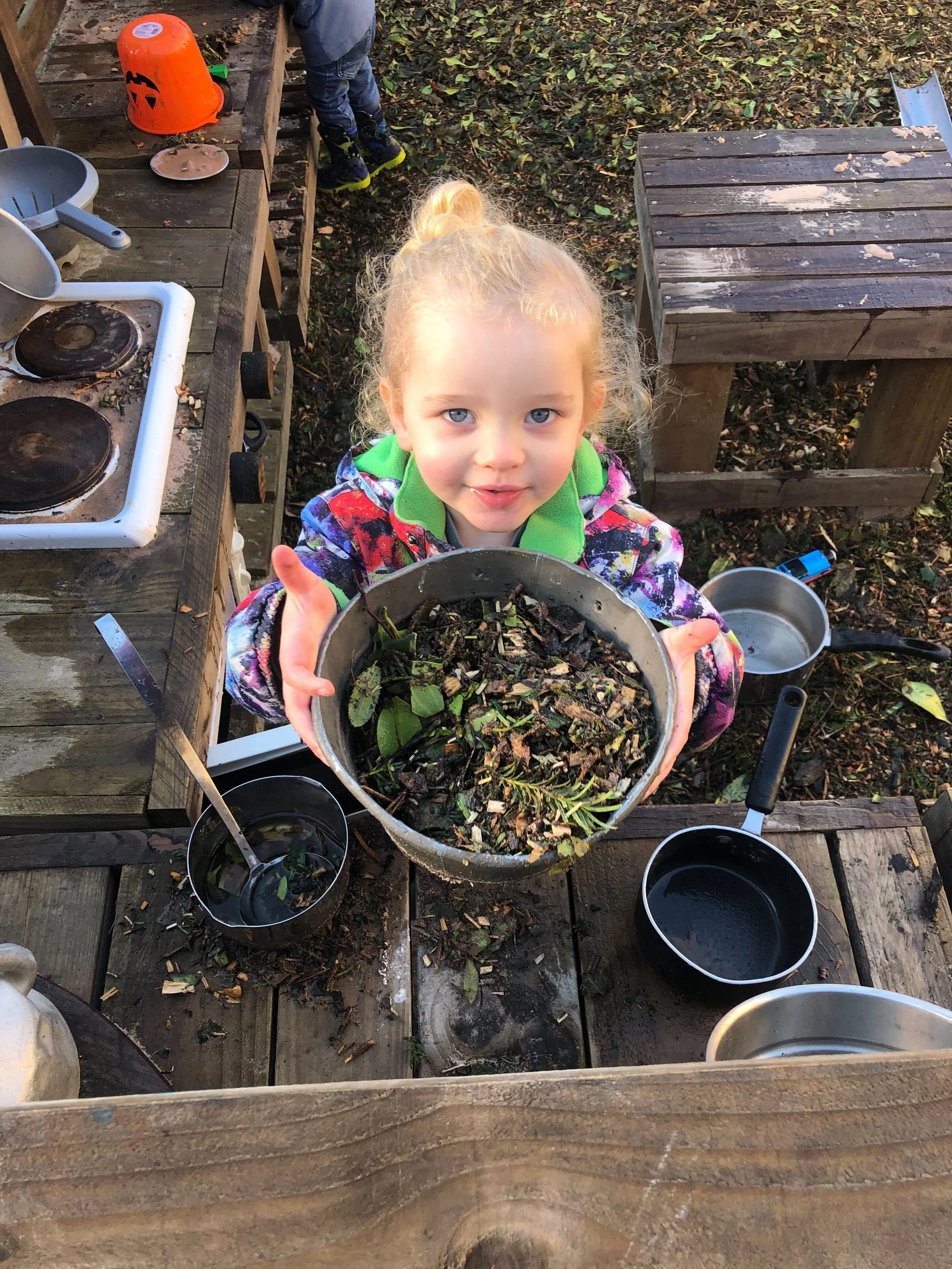 A young girl with blonde hair and blue eyes holding a metal bowl filled with leaves, twigs, and natural debris, standing outdoors on a wooden deck with scattered leaves, surrounded by various pots and kitchen tools.