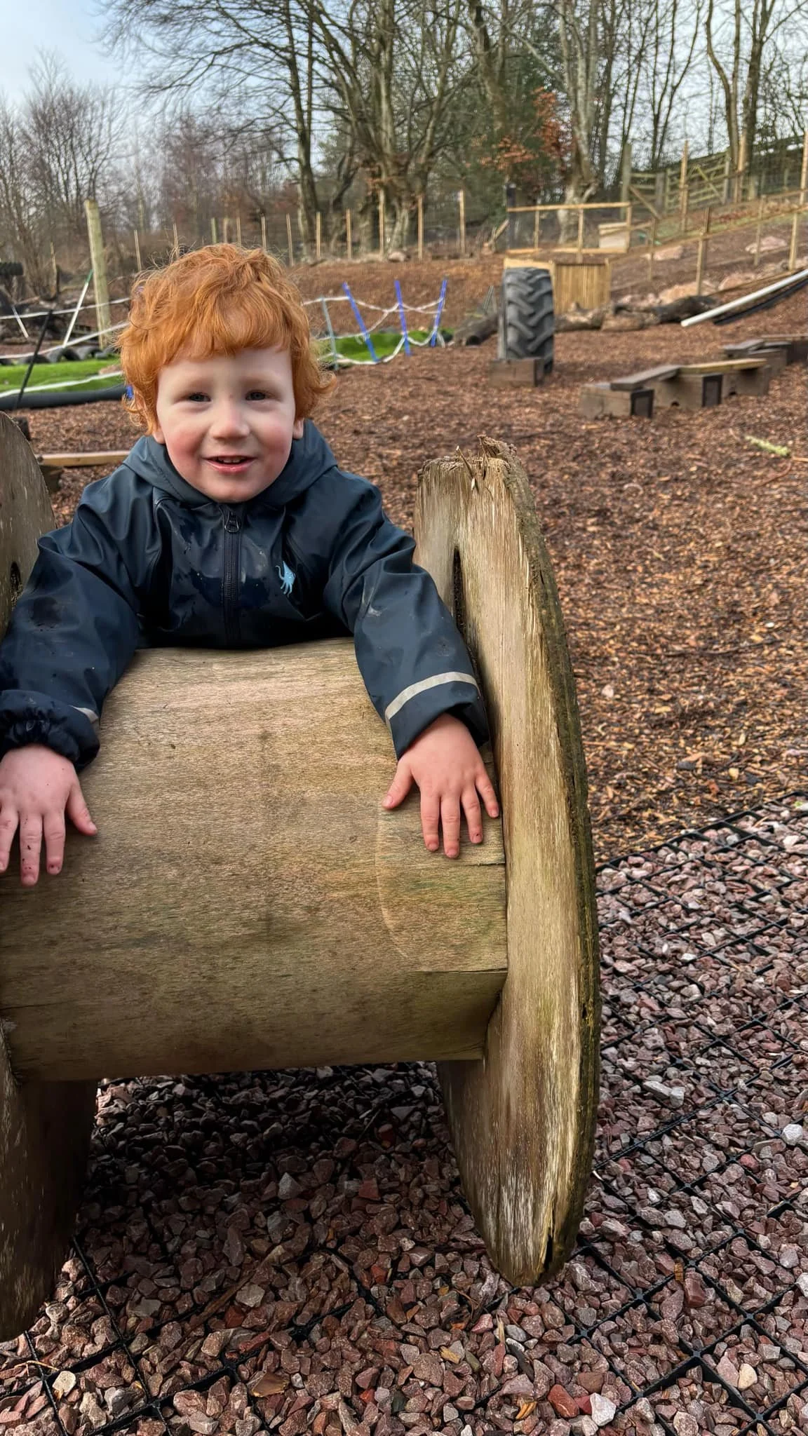 A young boy with red hair smiling while climbing on a wooden play structure at an outdoor playground during daytime. The background features trees and playground equipment.