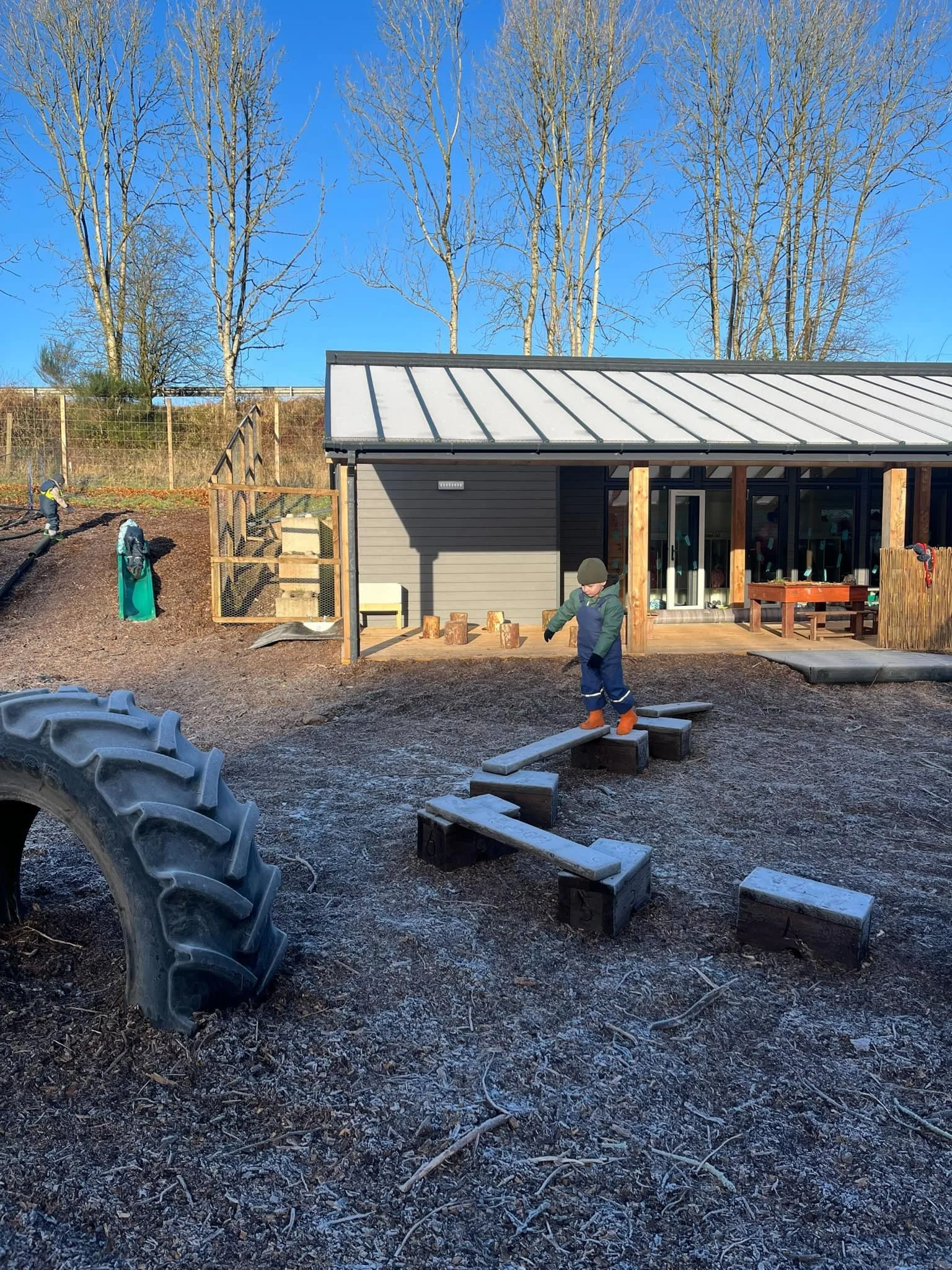 Children playing outdoors on a wooden deck in front of a modern building with large glass windows, surrounded by leafless trees and outdoor play equipment, under clear blue skies.
