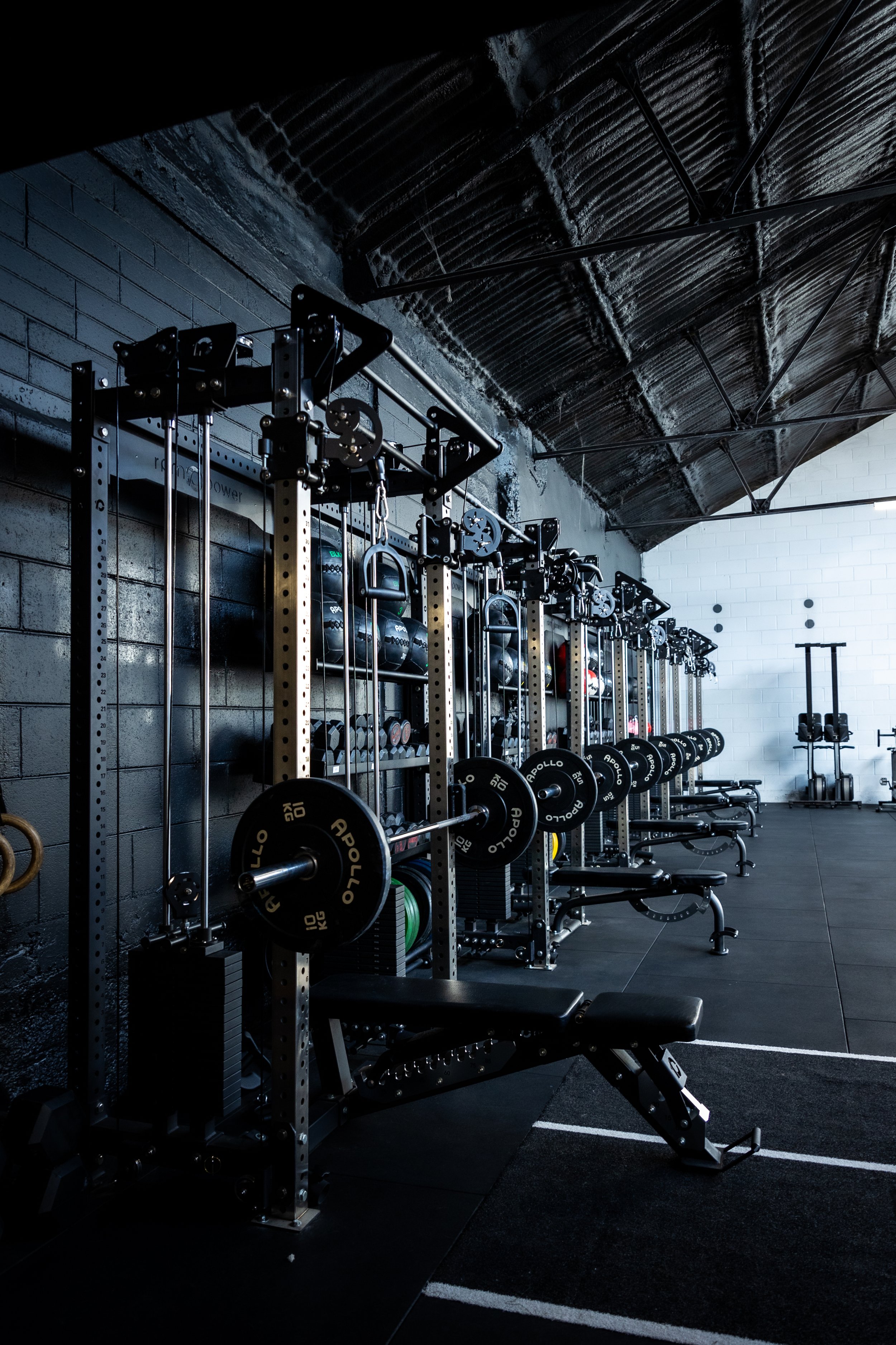 Row of gym weightlifting equipment, including barbells with black weights, benches, and racks in a dimly lit industrial-style gym with black and white walls.