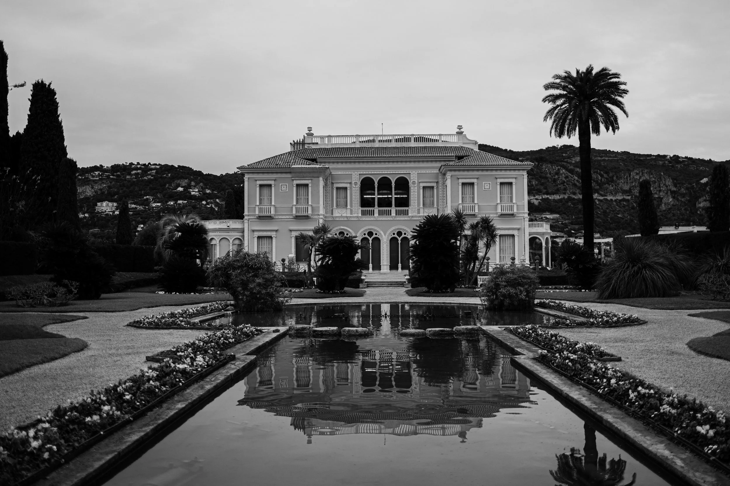 A large, ornate mansion with multiple windows and balconies, situated behind a landscaped garden with trees and bushes, reflected in a narrow pond in the foreground, with mountains in the background, all in black and white.