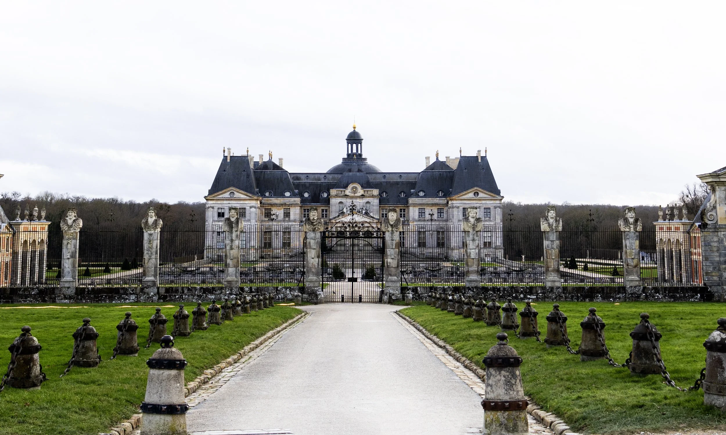 A grand historic castle with elaborate architecture, surrounded by a wrought iron fence and stone pillars with lion sculptures, accessed by a wide pathway flanked by green lawns.