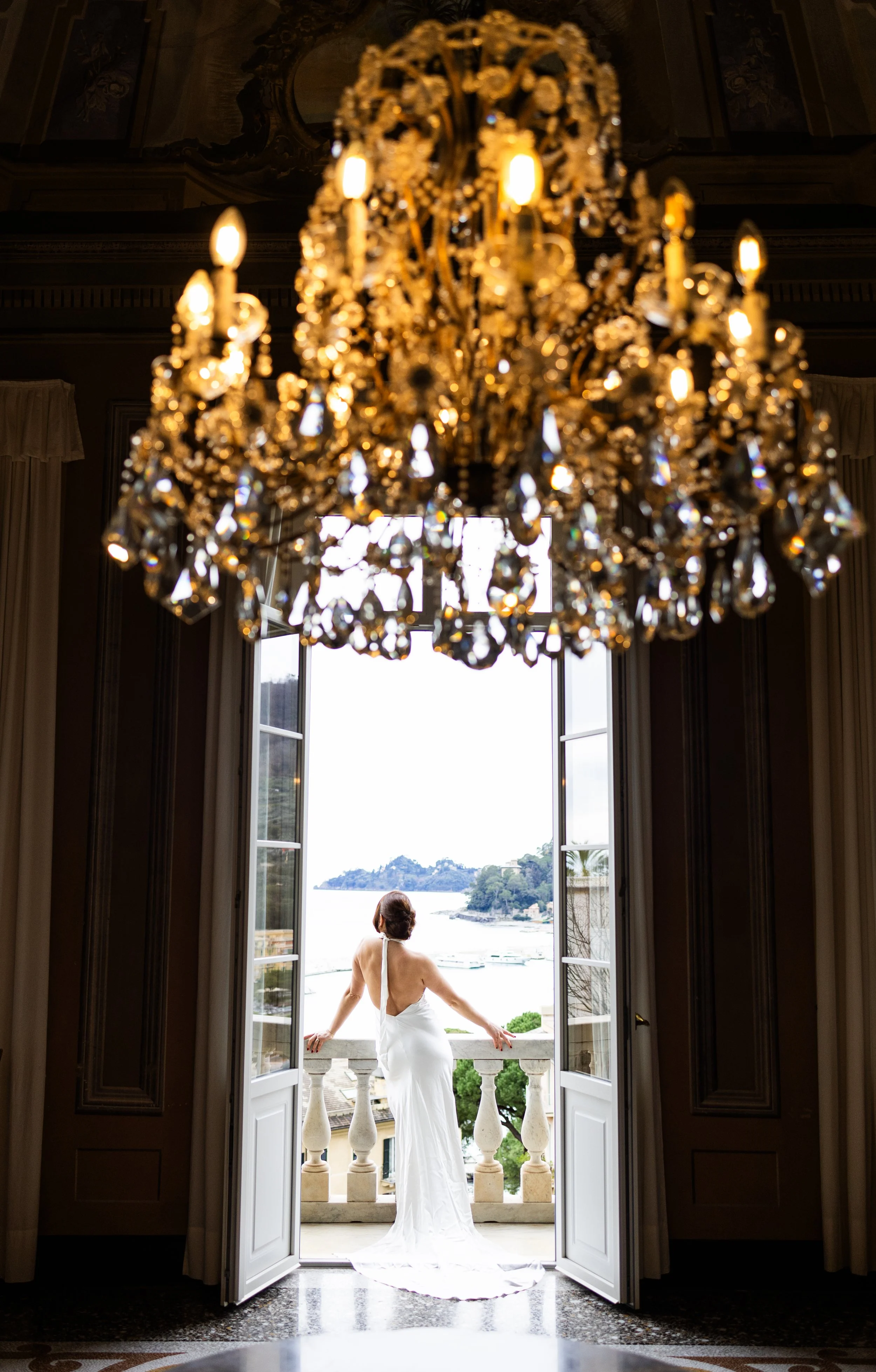 A woman in a white wedding gown standing on a balcony, gazing out over water and trees, seen through open double doors from a room with a large chandelier.