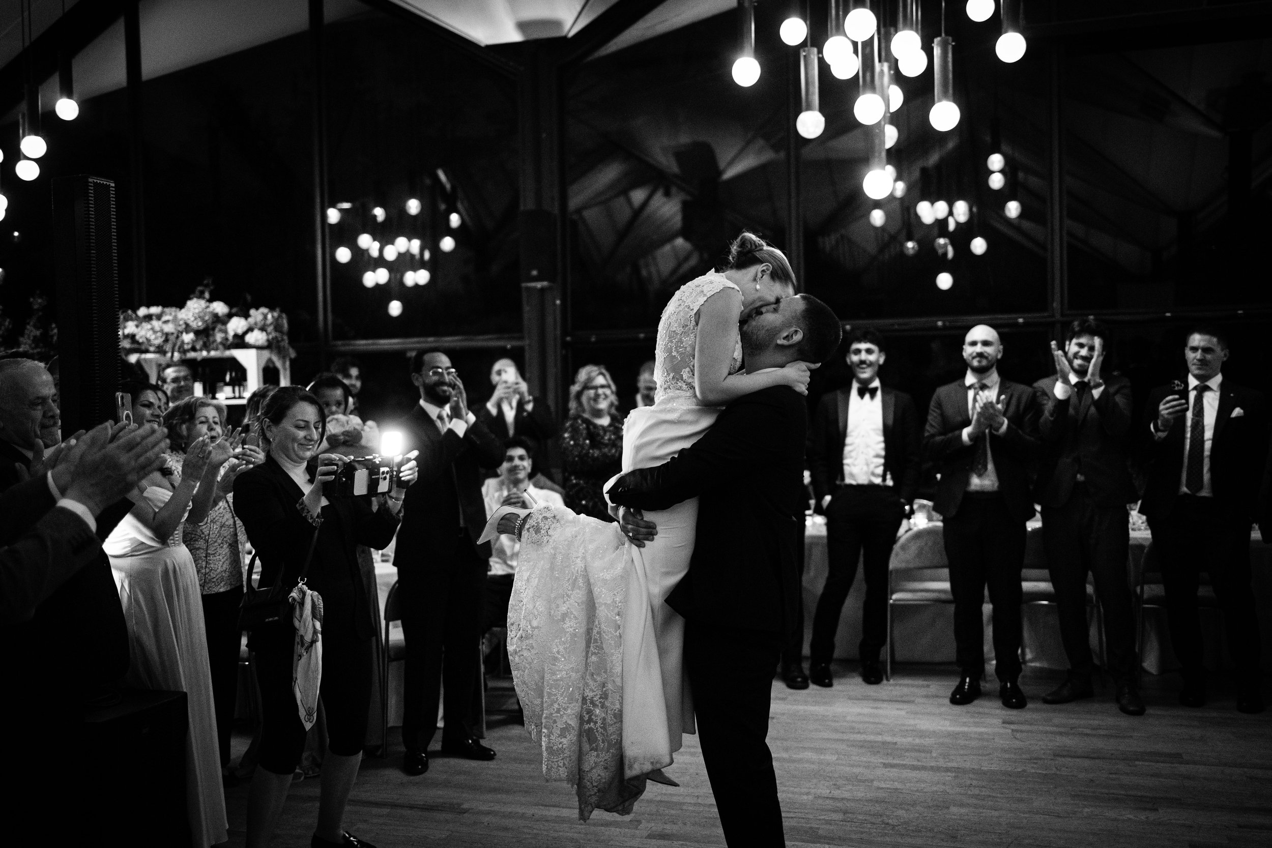 A bride and groom are dancing at their wedding reception while surrounded by guests clapping and taking photos. The bride is being lifted by the groom in a celebratory moment.