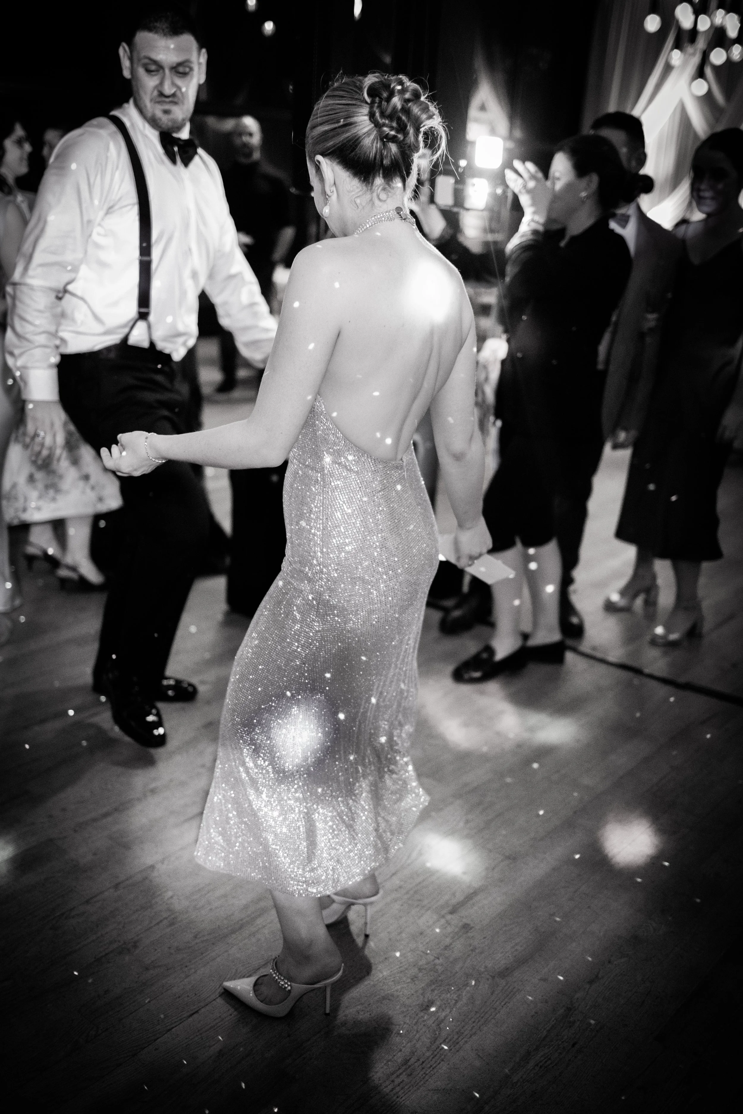 A woman in a backless, sparkling dress dancing at a party, surrounded by other guests in formal attire.