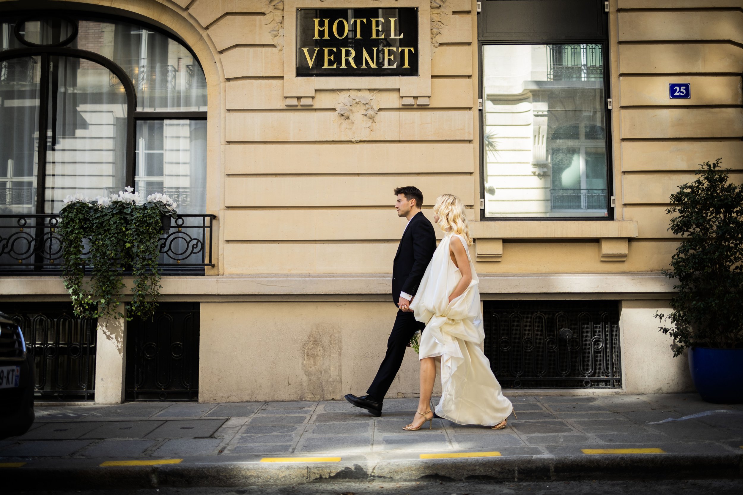 A man in a suit and a woman in a white dress walking on a city sidewalk in front of a beige building with a sign that says 'HOTEL VERNET'.