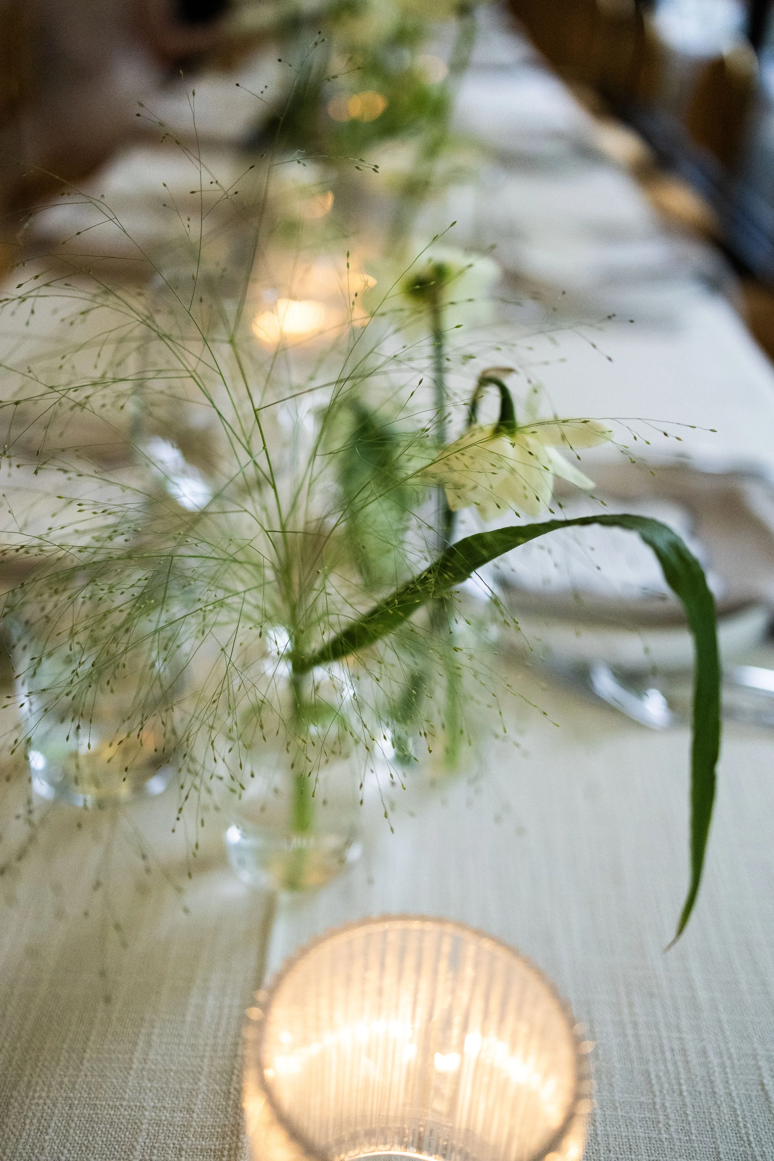 Close-up of a delicate floral arrangement with white flowers and green foliage in a glass vase, lit by a small candle on a table with a beige tablecloth.