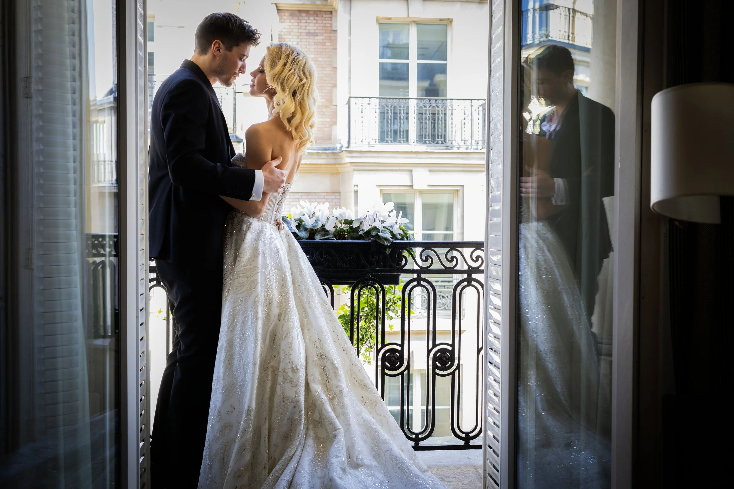 A bride and groom standing on a balcony, embracing each other. The bride wears a white wedding gown and the groom is in a black suit. Their reflection is visible in the glass door next to them.