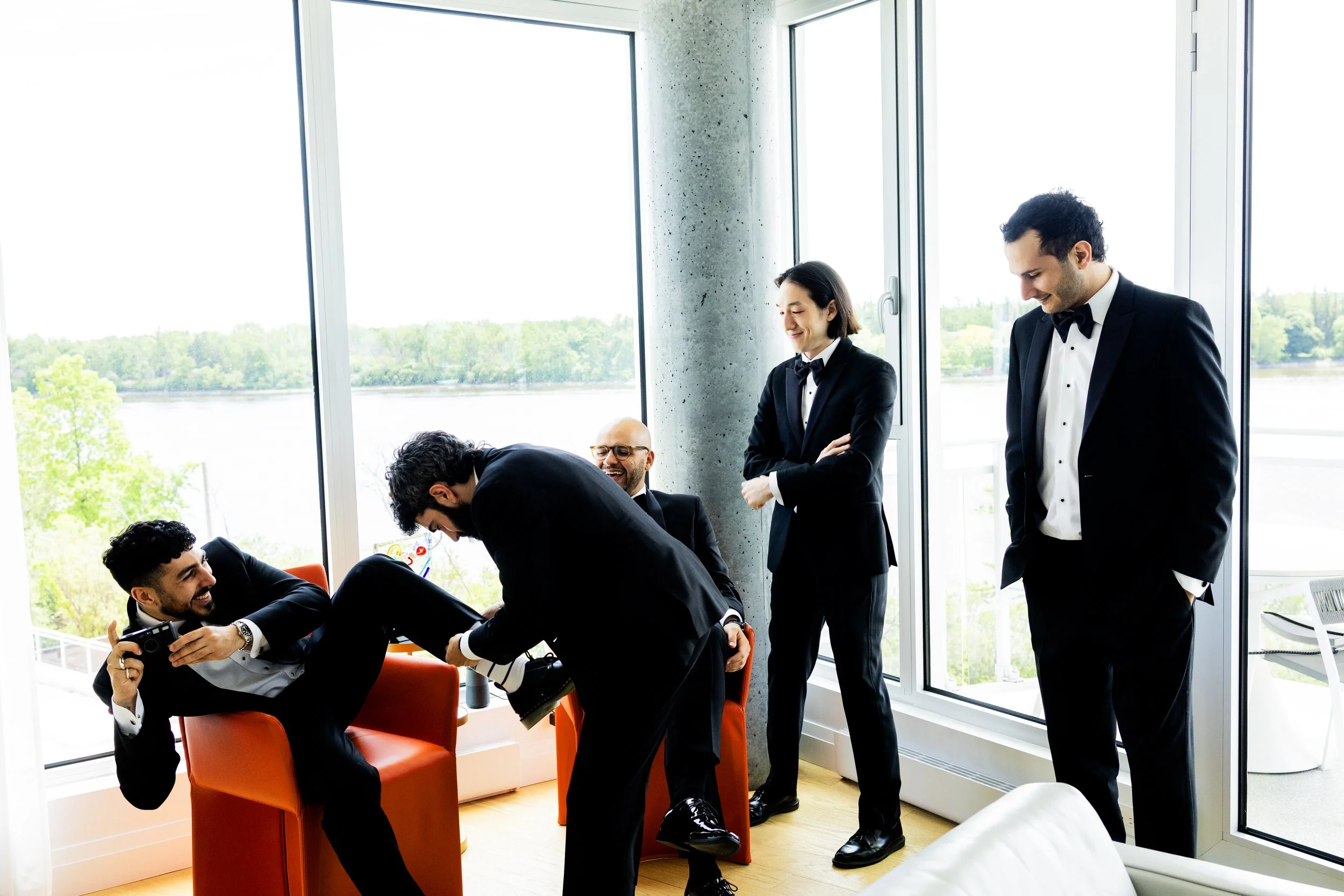 Group of five business professionals in formal suits having a fun moment while taking photos and laughing in a modern conference room with large windows overlooking a river and trees.