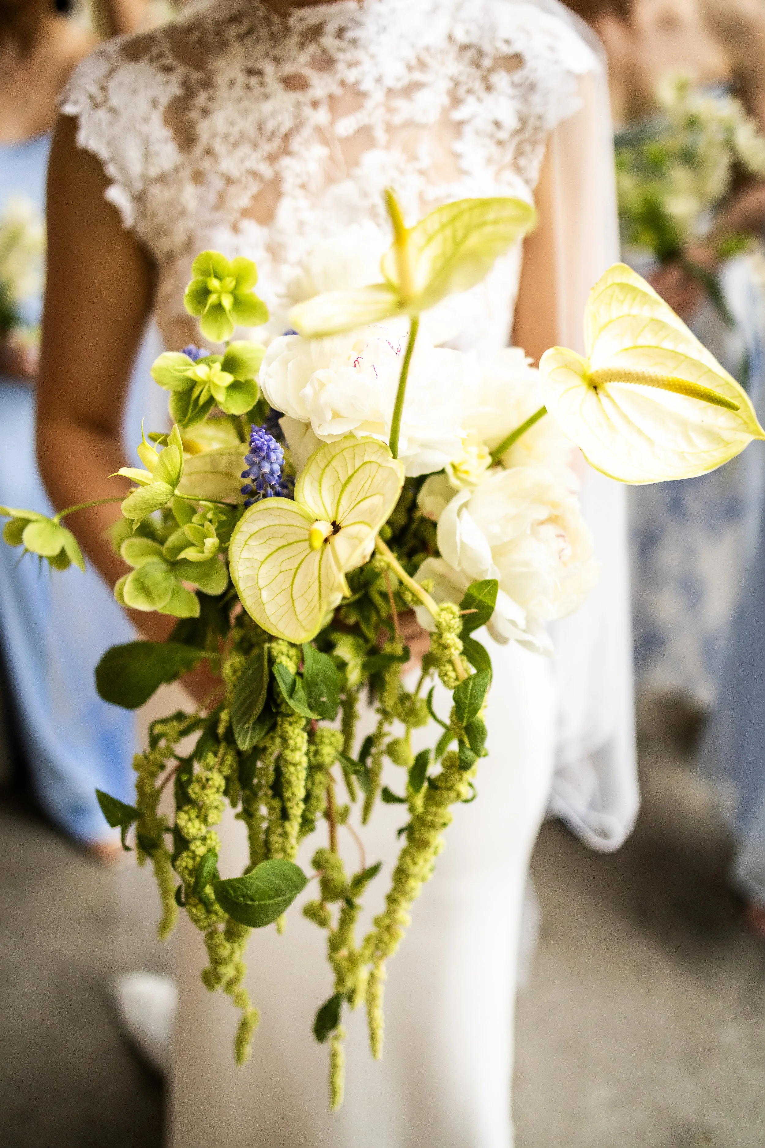 Close-up of a bride holding a cascading bouquet of white, green, and purple flowers, with a lace wedding dress partially visible.
