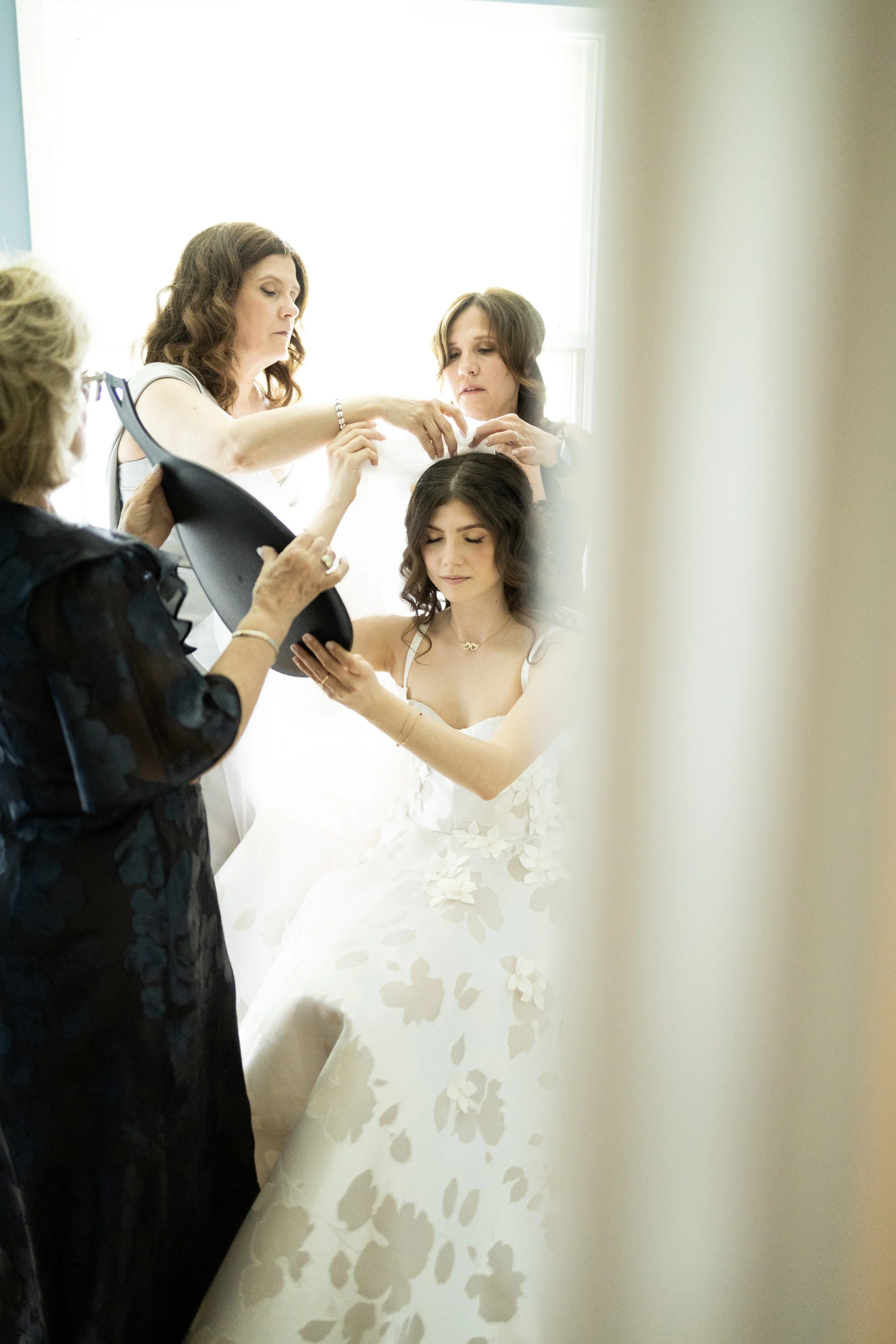 A bride sitting in a white wedding dress while women help her prepare, with one woman adjusting her hair and others assisting with her accessories in a bright room.