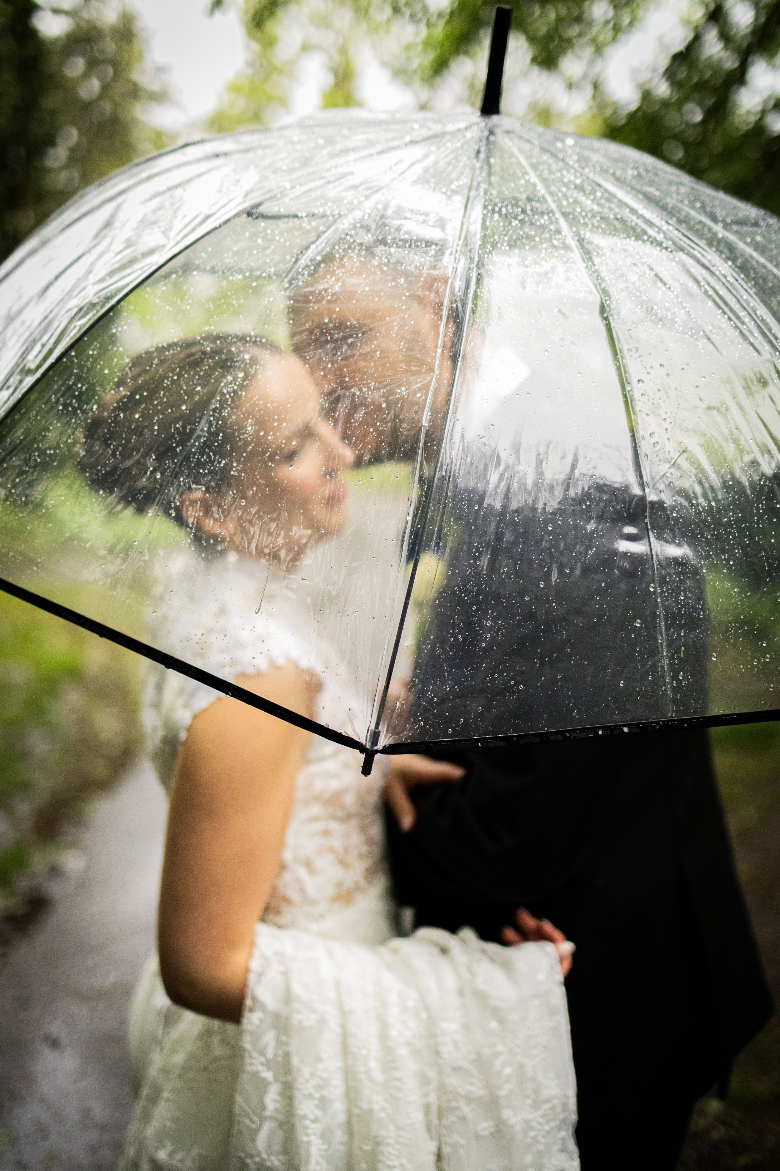 A couple sharing a kiss under a clear umbrella during rain, with lush greenery in the background.