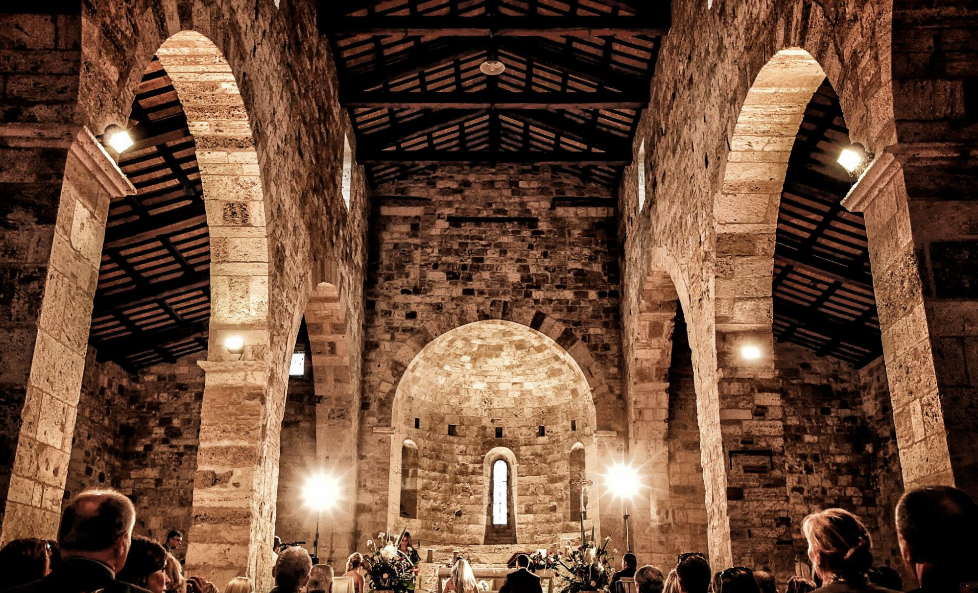 Interior of a stone church or chapel with arched windows, high vaulted ceiling, and congregation gathered for a ceremony or service, lit by spotlights and candles.