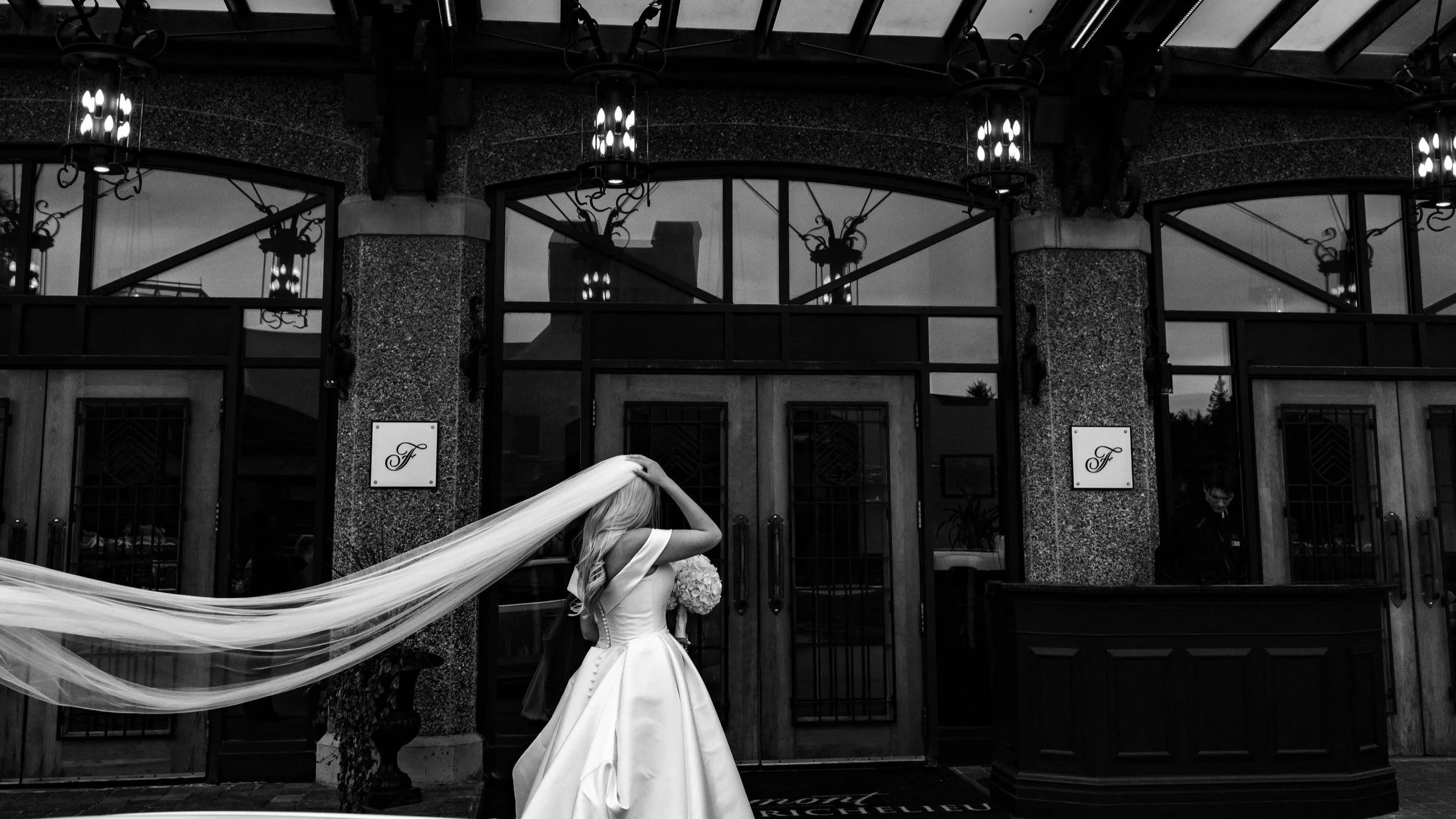 A bride in a wedding gown with a long veil standing outside a building, holding a bouquet of flowers, with a person in the background near the entrance.