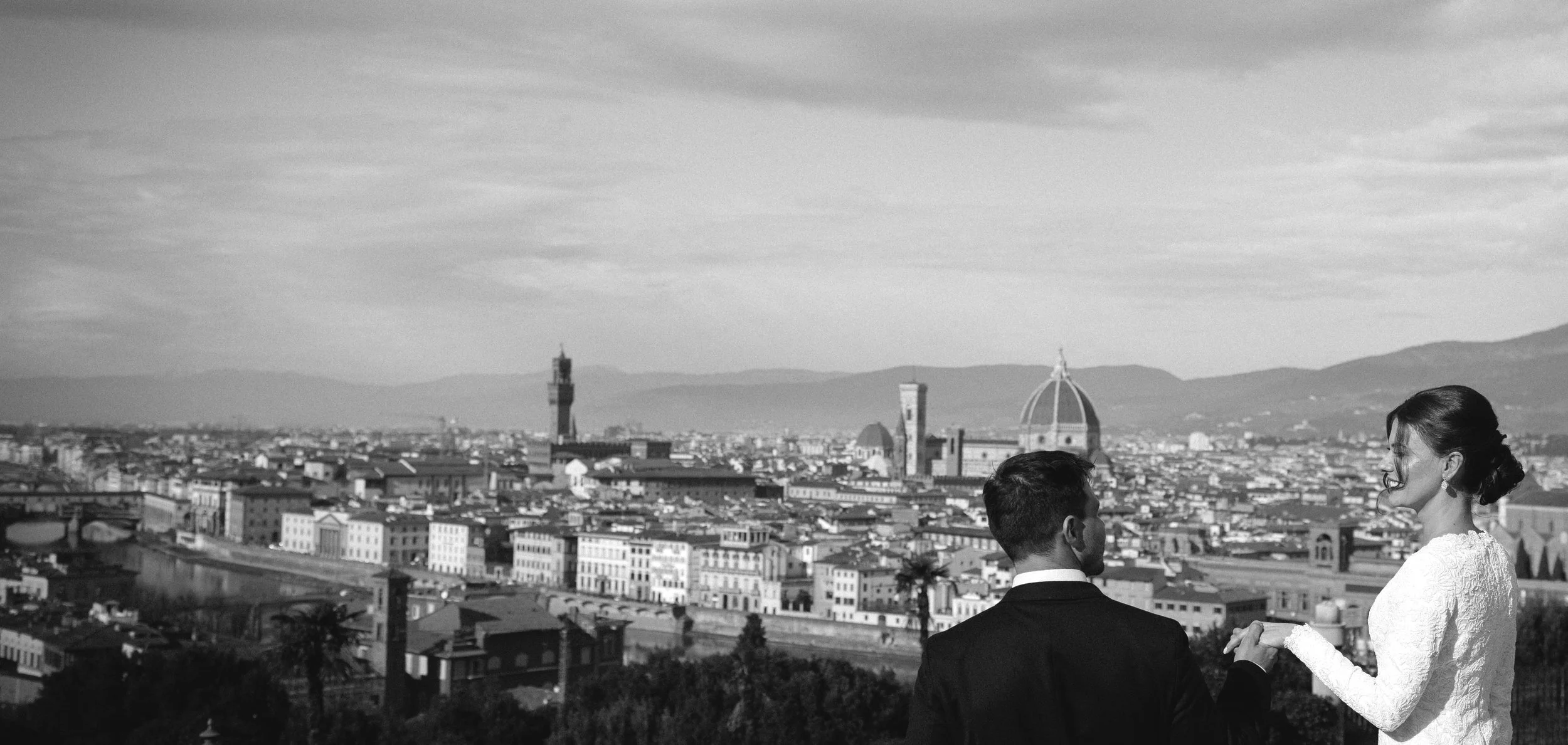 A black-and-white photo of a cityscape with a couple holding hands, facing each other, with the skyline and mountains in the background.