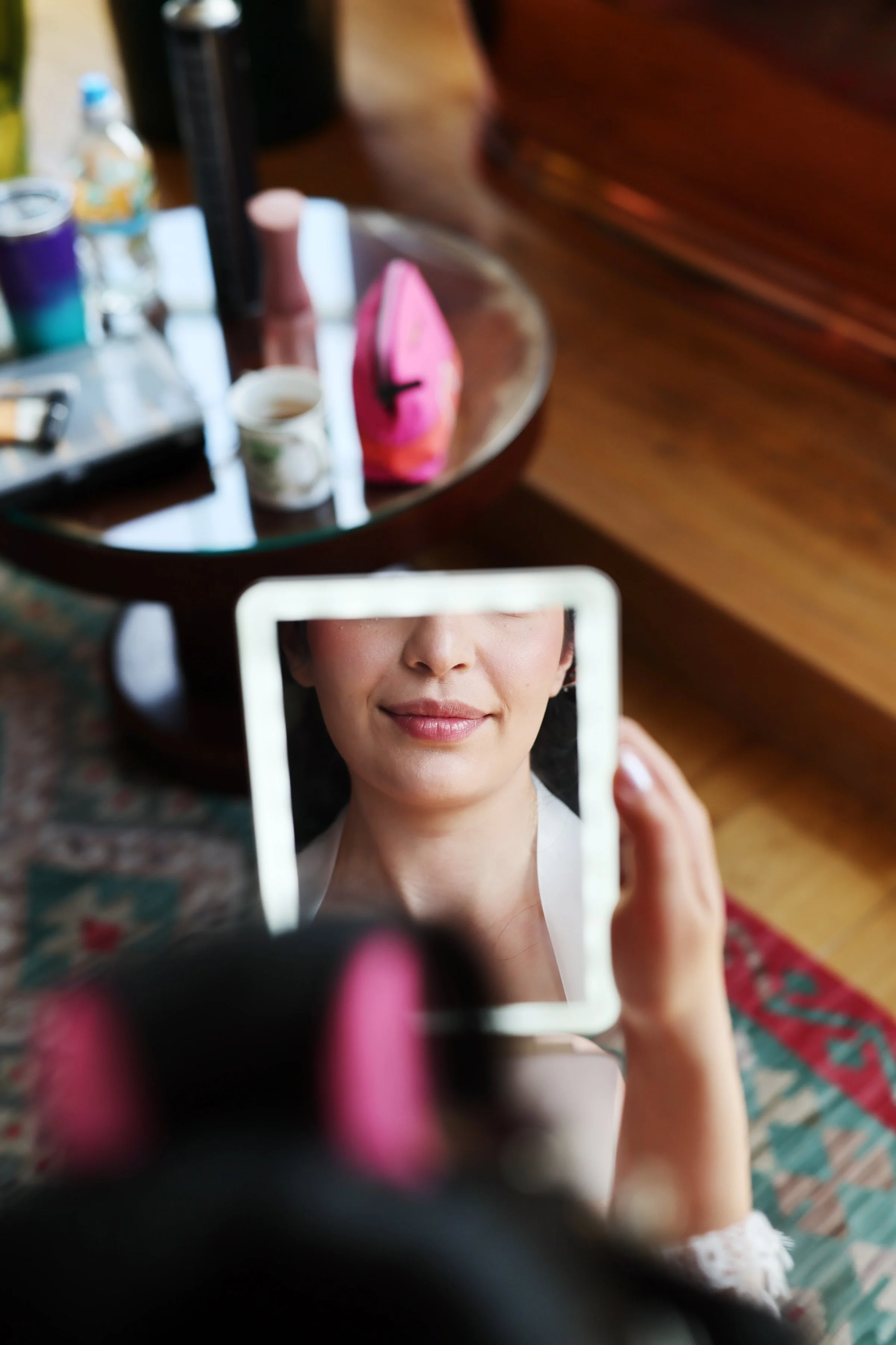 A woman holding a small mirror, capturing her face with a smile, in a room with a table that has various cosmetic items and bottles on it.