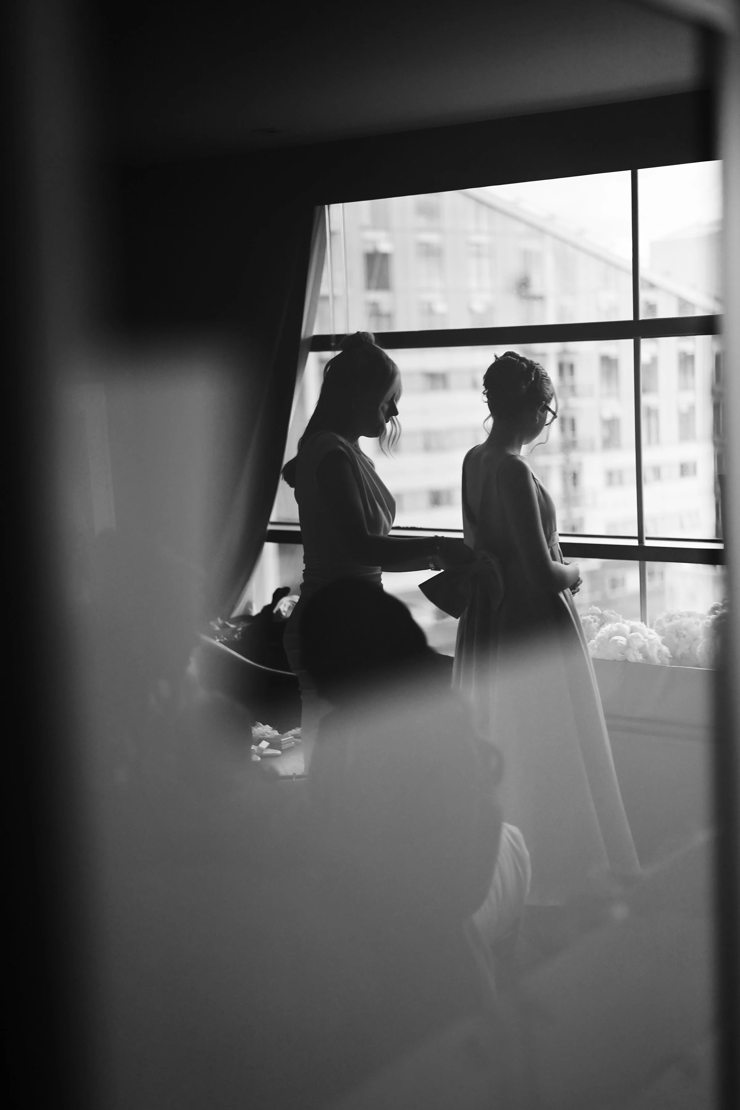 Black and white photo of two women standing near a large window. One woman is adjusting the other's dress. The scene appears to be indoors with a cityscape in the background.