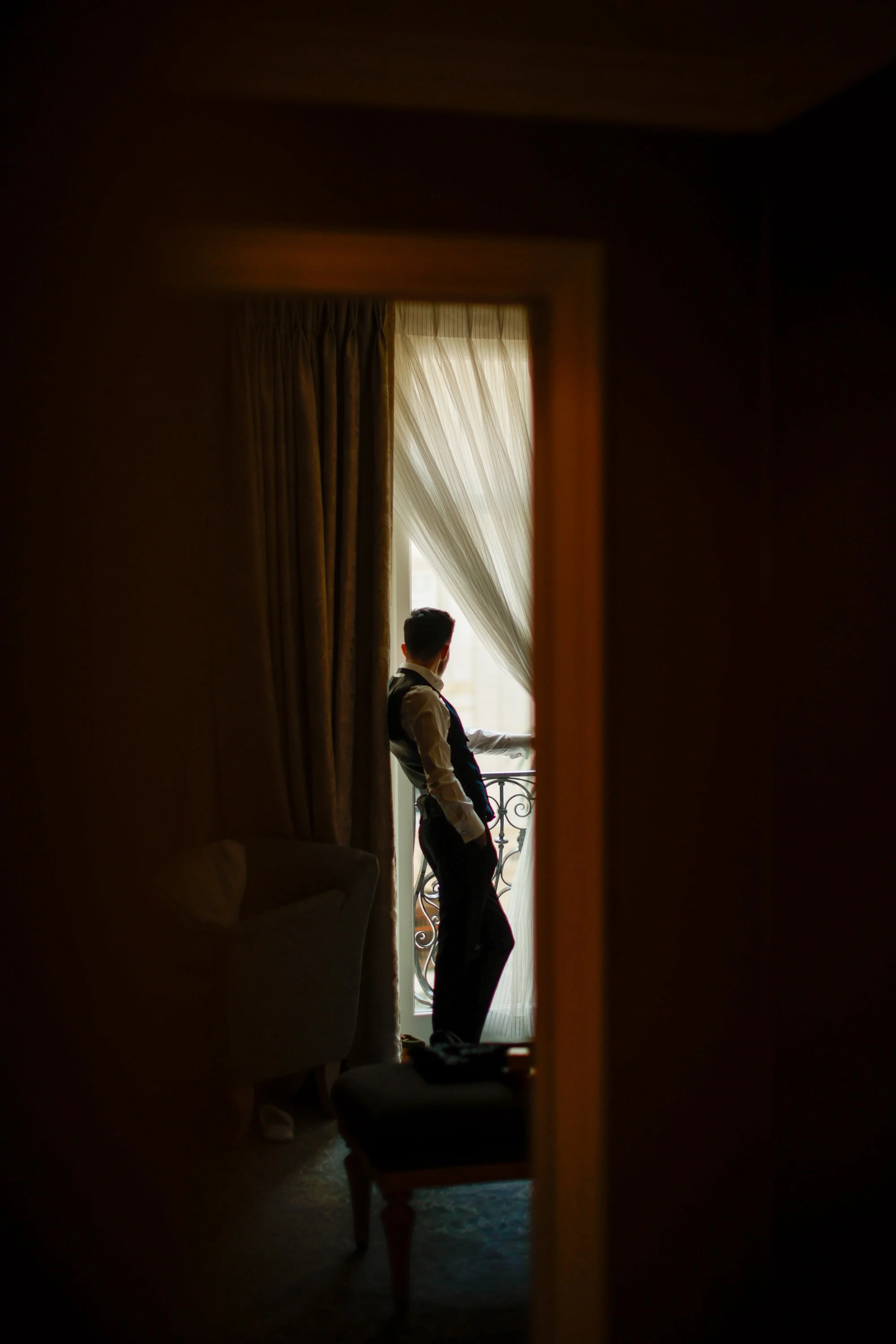 A man in formal attire is standing by a window with sheer curtains, leaning against a balcony railing and looking outside, viewed through a mirror in a dimly lit room.